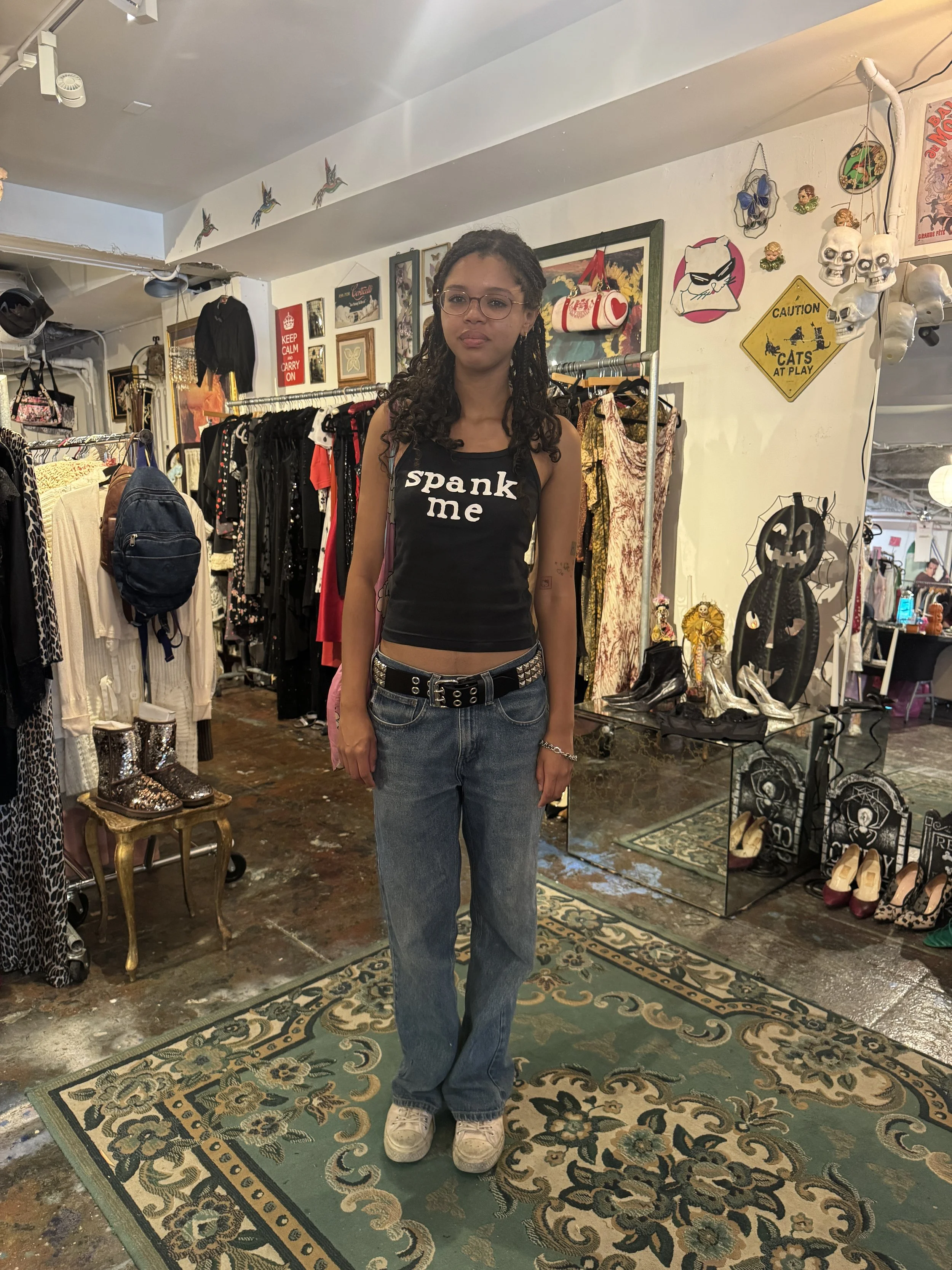 Young woman with glasses and dreadlocks standing in a vintage clothing store, wearing a black crop top with the words 'spank me', blue jeans, and white sneakers.
