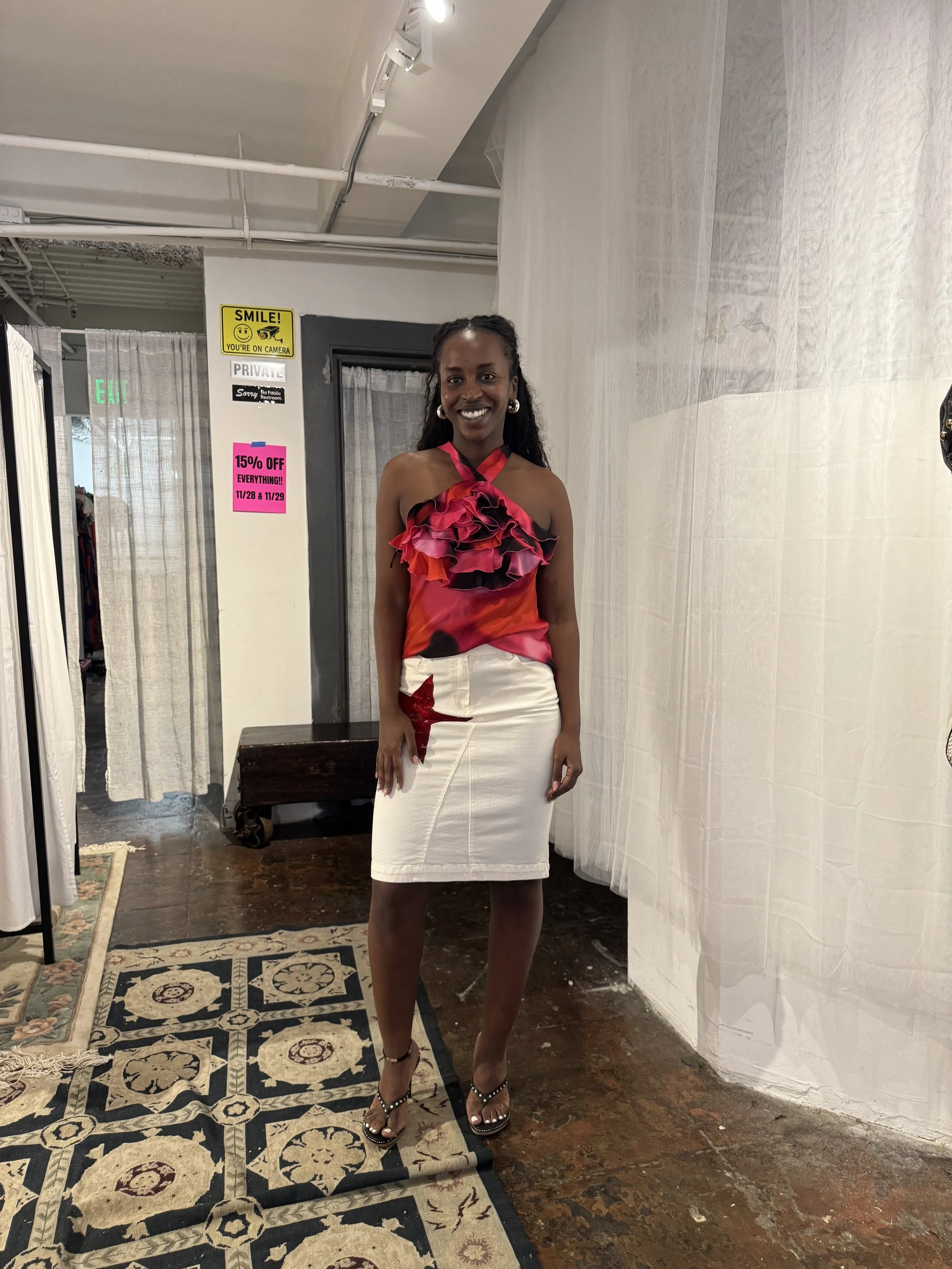 A woman standing in a boutique wearing a colorful ruffled top and a white skirt with red star details, smiling at the camera.