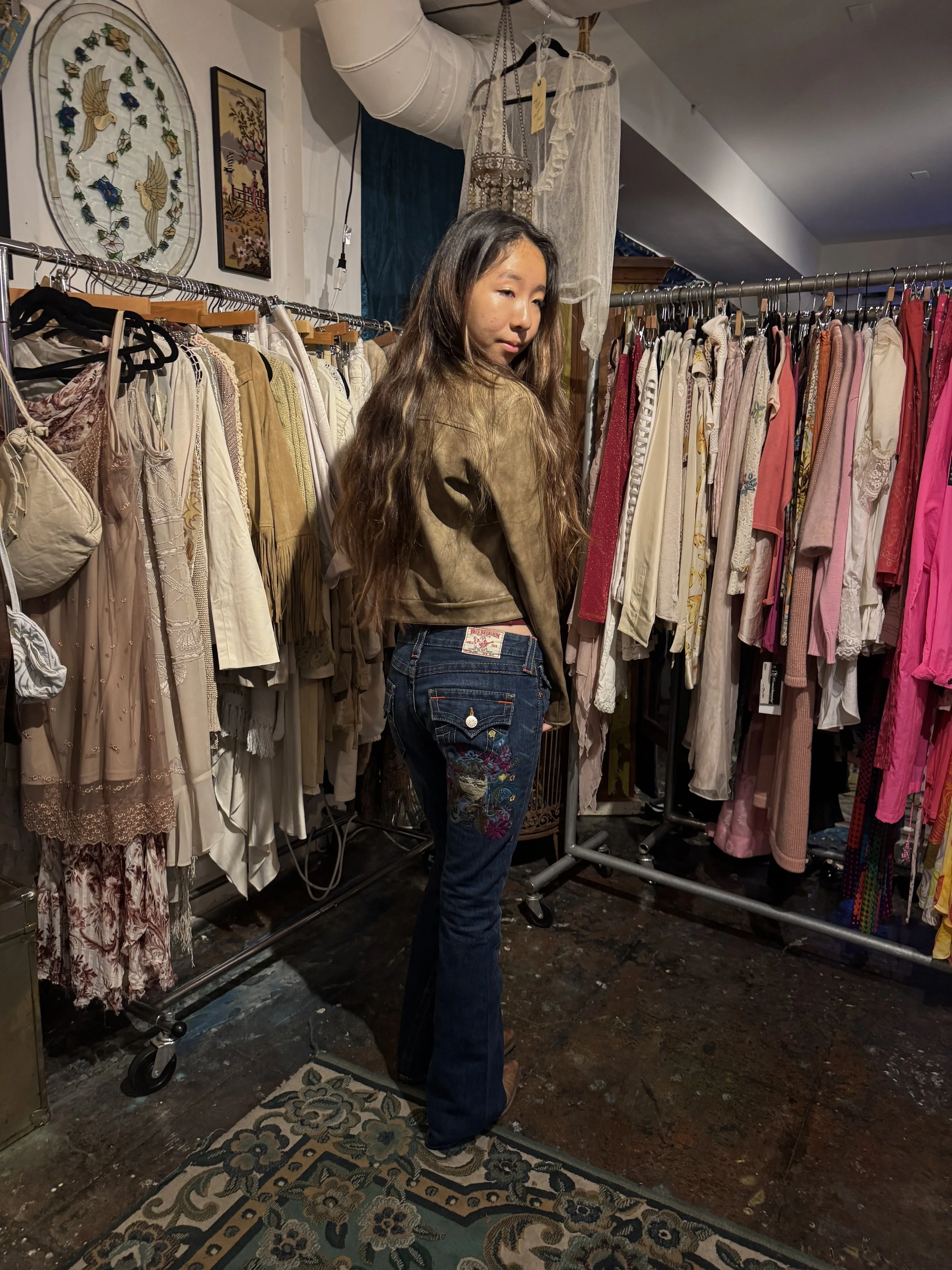 A woman with long wavy hair looking over her shoulder in a vintage clothing store, surrounded by racks of dresses, blouses, and other apparel.