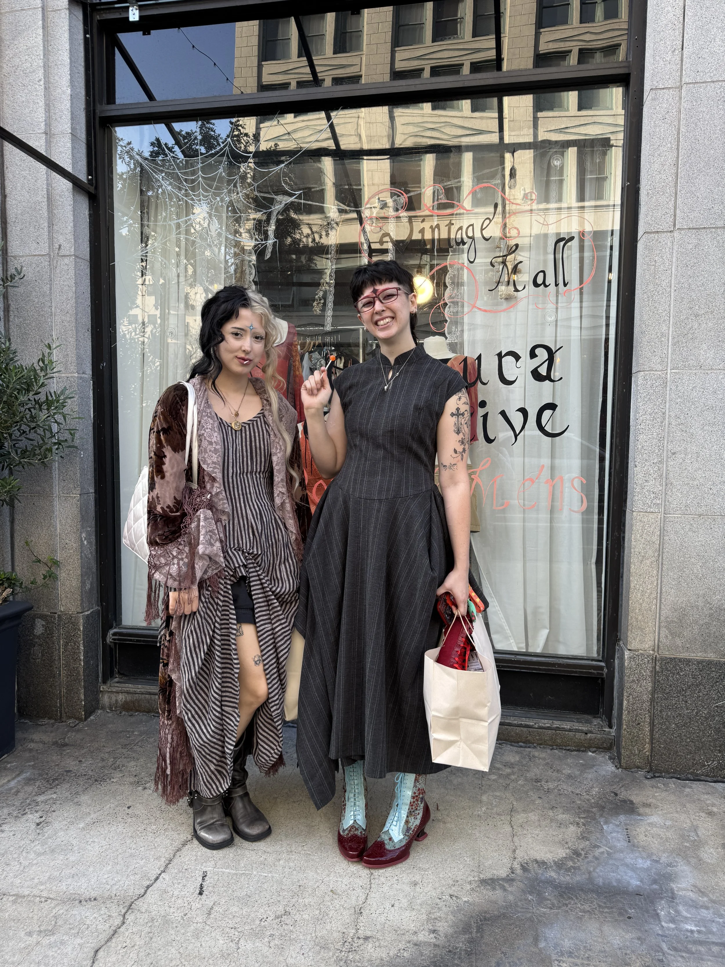 Two women with unique, eclectic fashion standing outside a boutique with a glass window display. One woman has long dark and blonde hair, wearing a striped pajama-like outfit with a long cardigan and combat boots. The other woman has short dark hair,