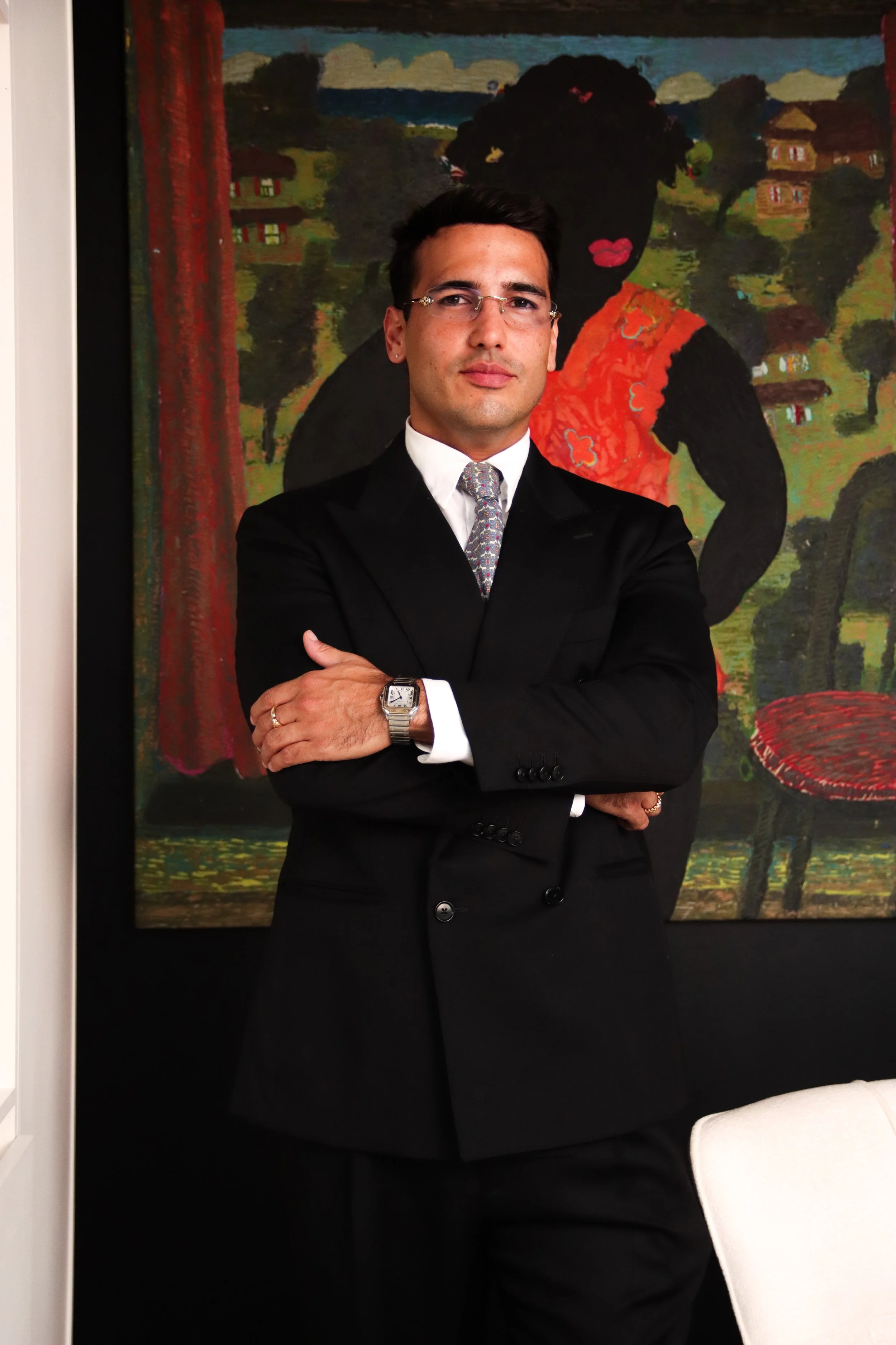 A man in a black suit and tie with glasses and a wristwatch, standing with crossed arms in front of a colorful painting of a woman with dark hair, red lips, wearing a red top, with small houses and trees in the background.