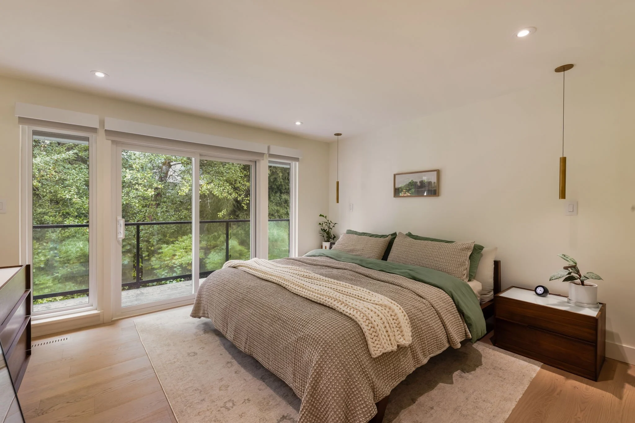 Bedroom with a large bed, sliding glass door leading to a balcony, view of trees outside, bedside table with plant and clock, minimalist decor, soft lighting.