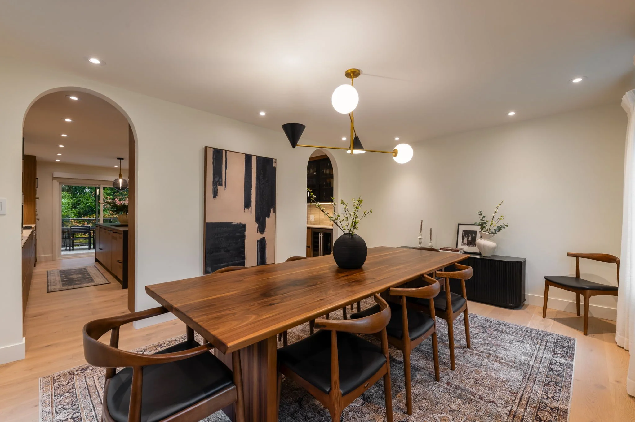 Dining room with wooden table, six chairs with black cushions, modern chandelier, abstract wall art, and decorative vases with greenery.