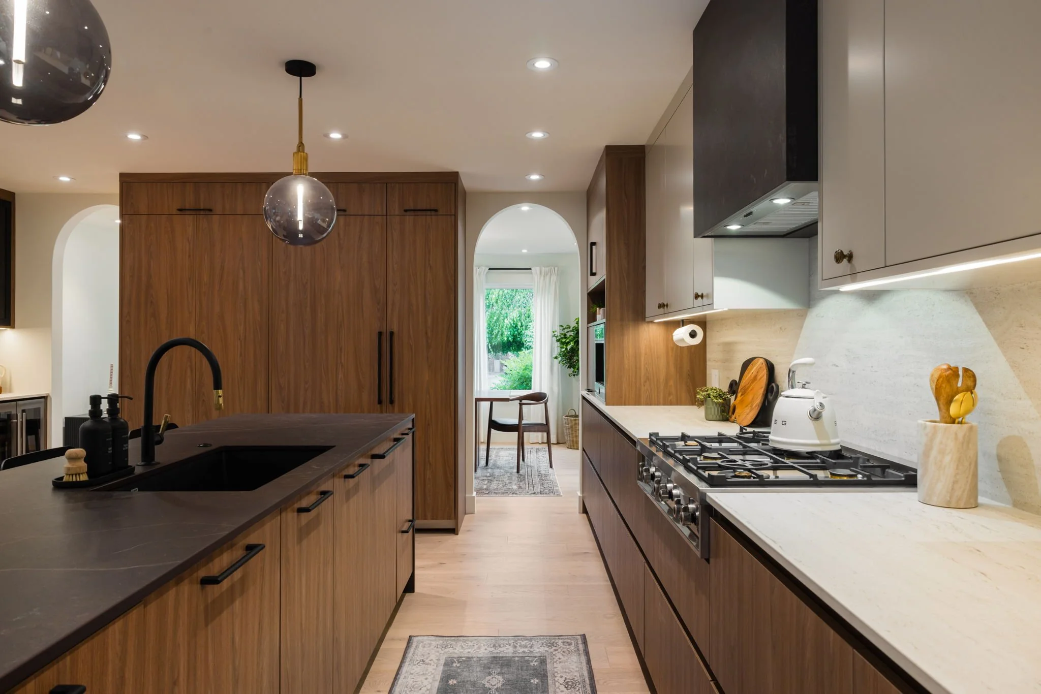 Modern kitchen with wooden cabinets, black countertops, and a white countertop with a gas stove. Pendant lights hang from the ceiling, and a small dining area with a window is visible in the background.