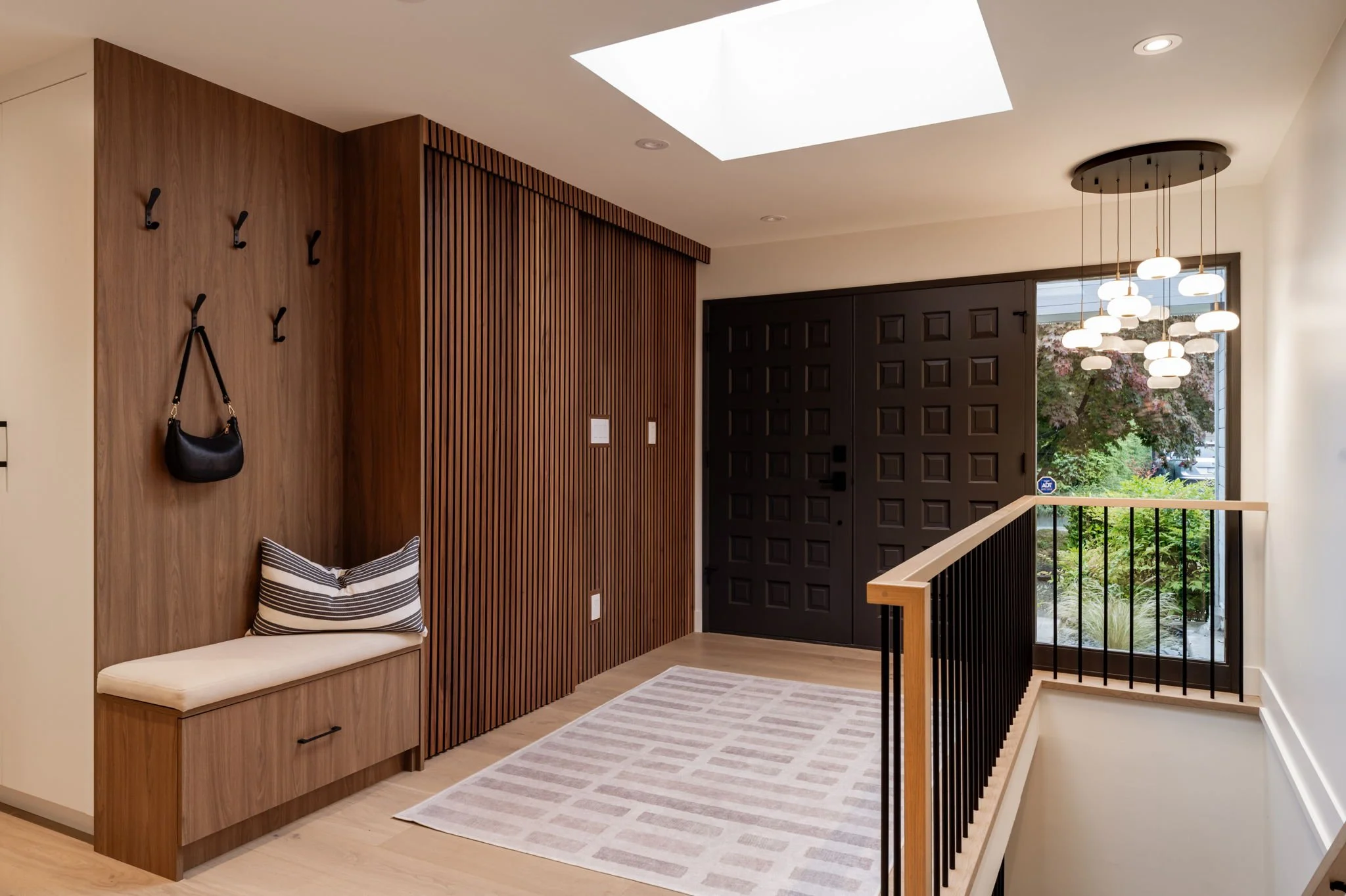 Modern entryway with a black front door, wooden wall paneling, a bench with a striped cushion, hooks with a handbag, a white and beige rectangular rug, a glass window showing outdoor greenery, and a contemporary hanging light fixture.