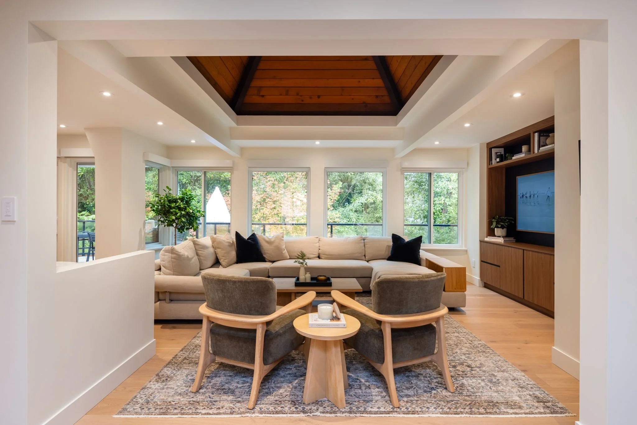 Modern living room with a beige sectional sofa, two grey armchairs, a small round wooden table, large windows with greenery outside, a wooden built-in TV cabinet, potted plants, a patterned area rug, and a wooden ceiling with a skylight.