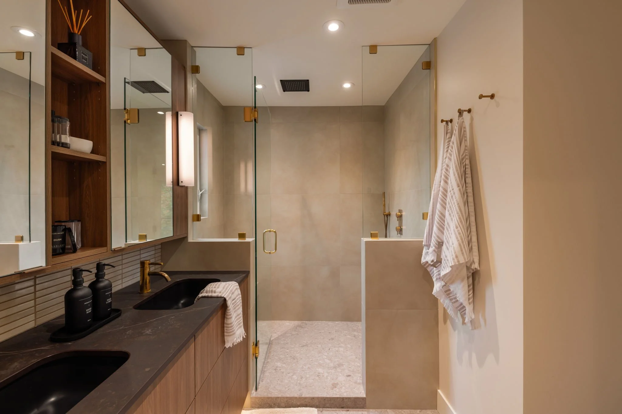 Modern bathroom featuring dual sinks with black countertops, gold fixtures, wooden cabinetry, and a glass shower enclosure with beige tile walls and a partial view of a bathtub.
