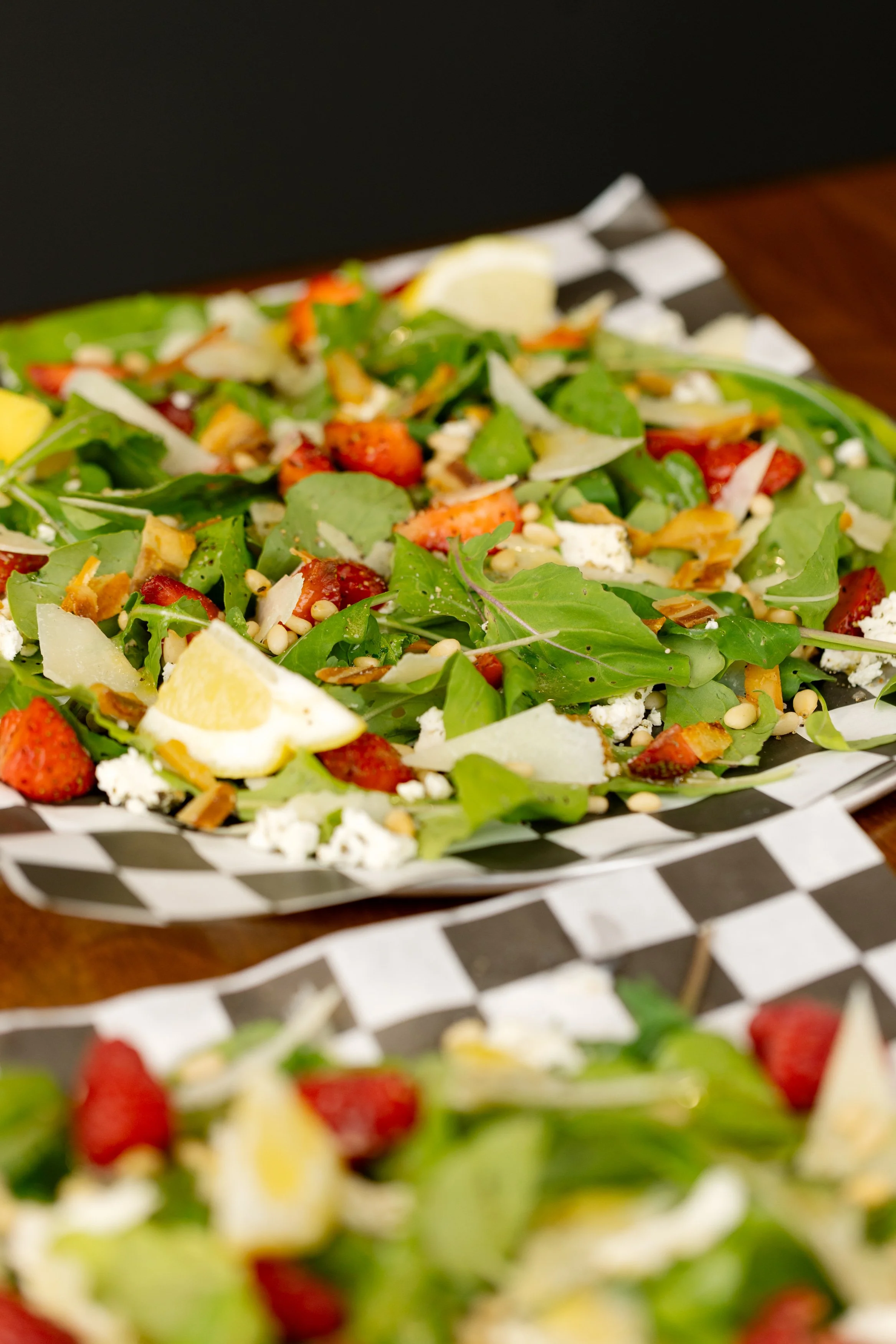 Fresh salad with mixed greens, strawberries, shredded cheese, and sliced vegetables in a black and white checkered paper tray.
