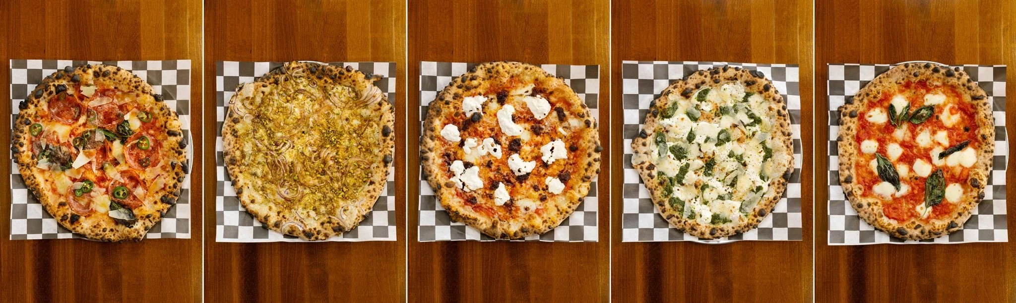Three pizzas with various toppings on a checkered pizza paper, placed on a wooden table.