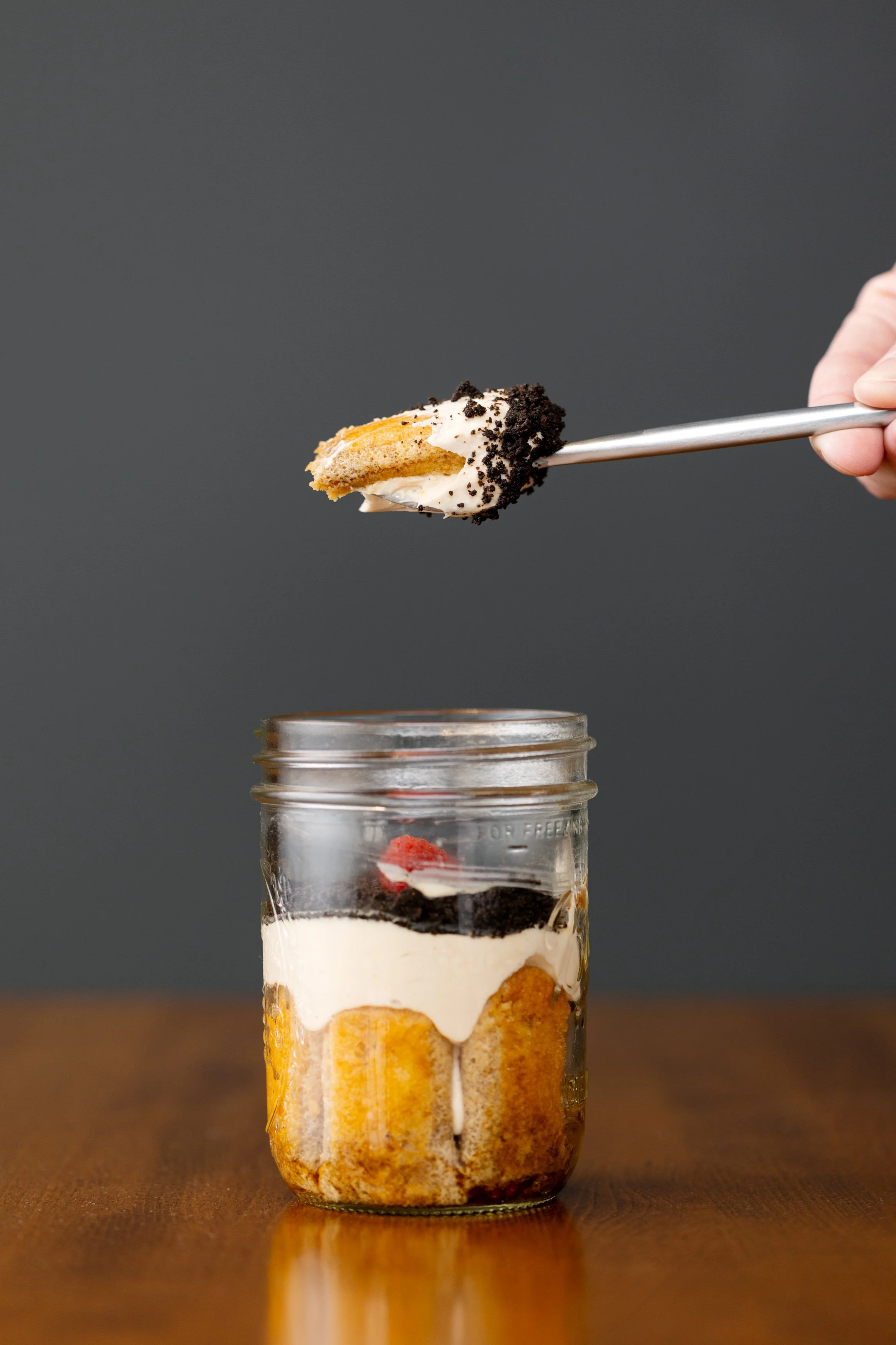 A hand holding a spoon with cake and cookie crumbs, over a Mason jar filled with layered cake, cookies, and cream, on a wooden surface.
