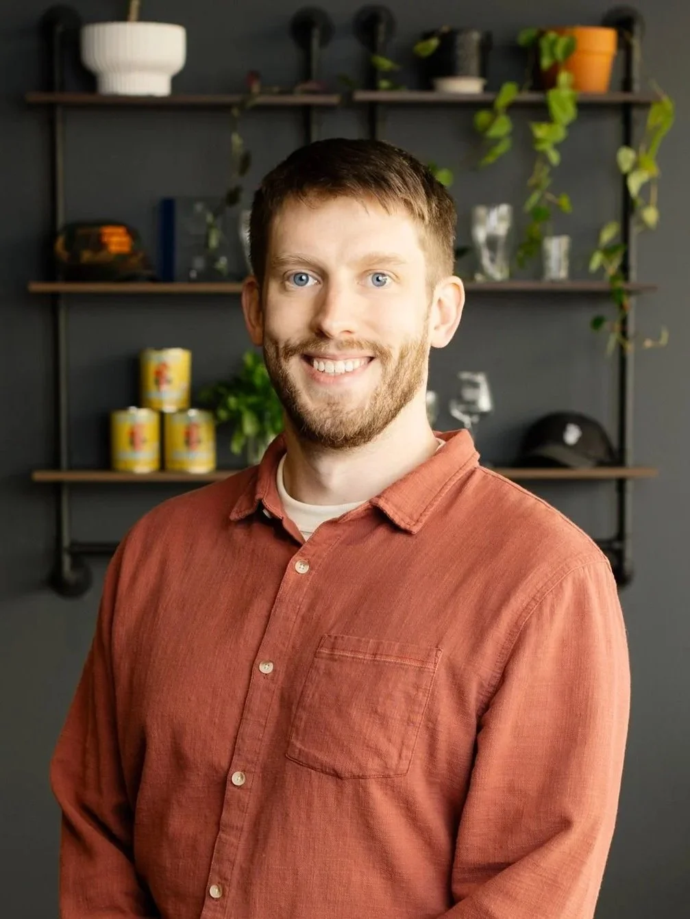 A young man with short brown hair, a beard, and blue eyes smiling, wearing a dark hoodie with a graphic design, standing in front of two blurry posters of coffee cups.