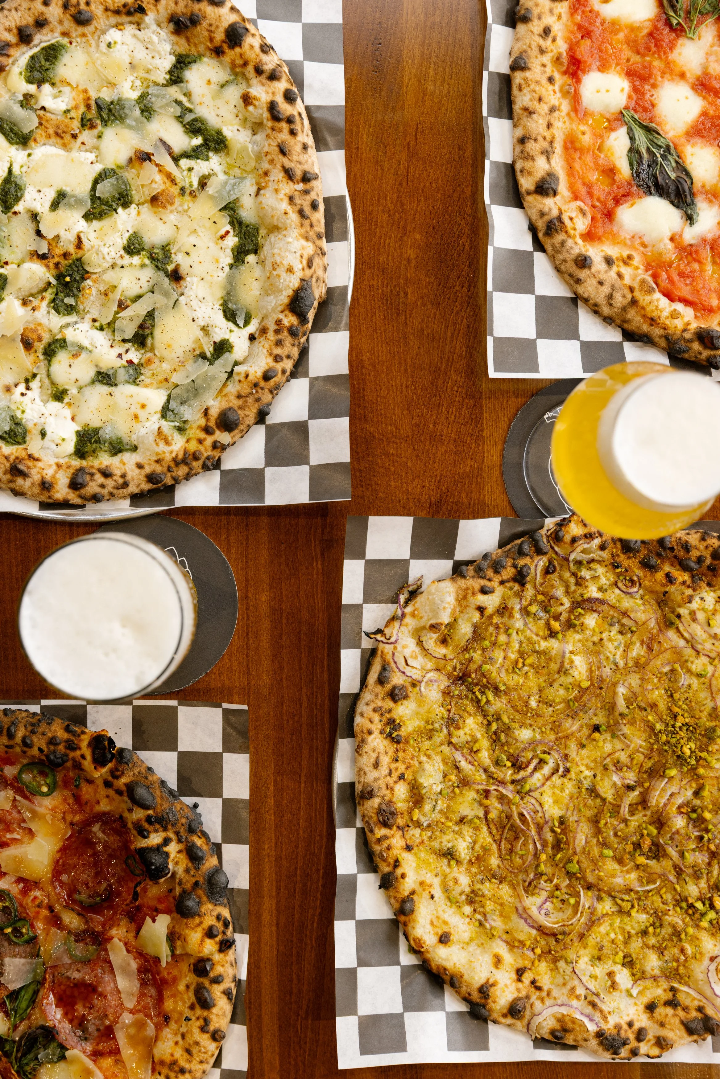 Several pizzas with various toppings on a checkered paper-lined tray on a wooden table, with glasses of beer beside them.