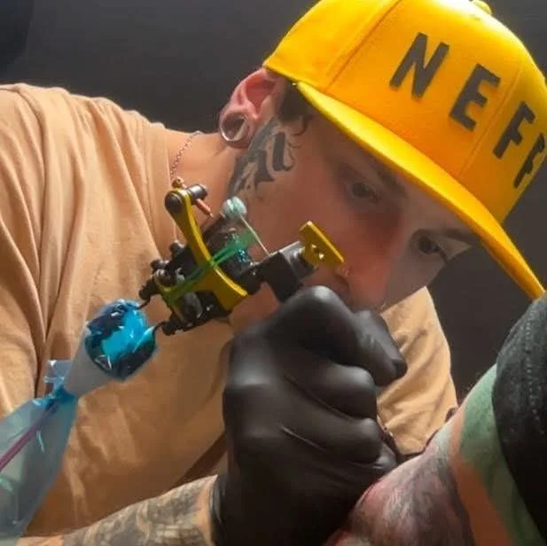 Tattoo artist in a brown shirt and yellow cap with black lettering, wearing black gloves, using tattoo equipment on someone.