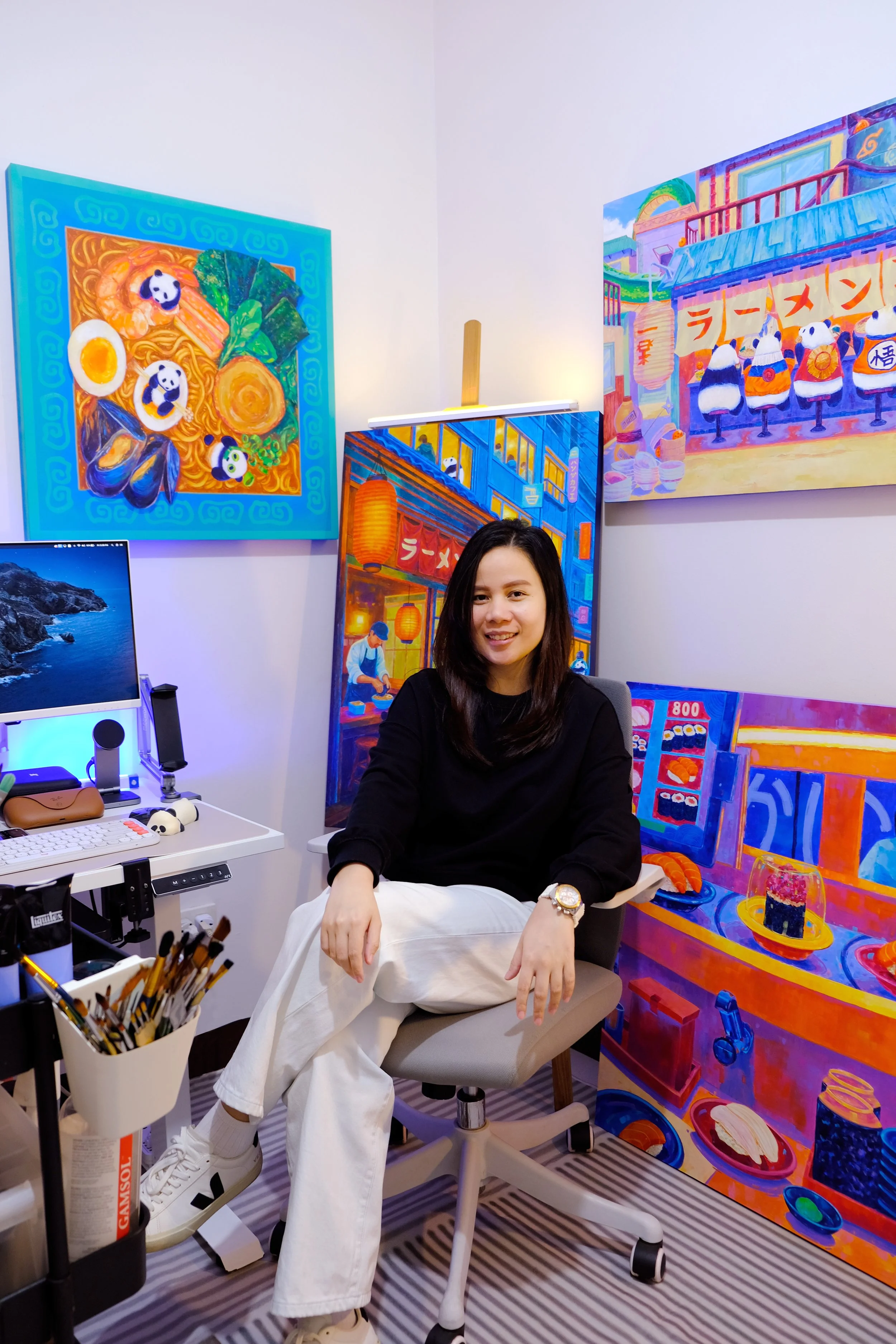 Portrait of the artist, Krissie Phee (kgphee) seated in their studio, surrounded by vibrant colorful paintings on the wall behind them.