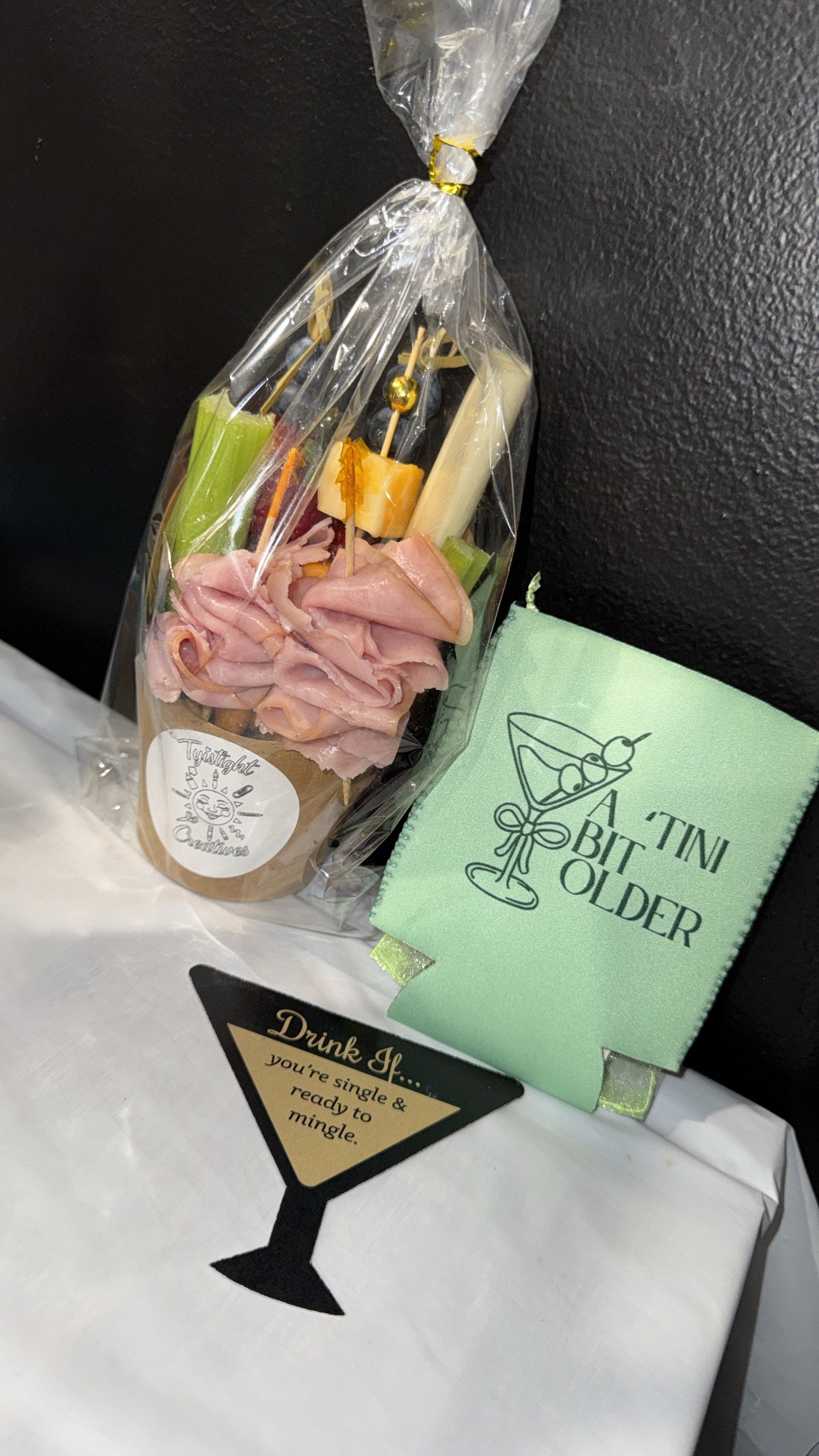 A clear plastic favor bag with assorted cheese, grapes, and sliced meats on a beige paper holder, with a green napkin that has 'Tini Bit O'Old' printed on it, along with a black paper drink tag on a white tablecloth.