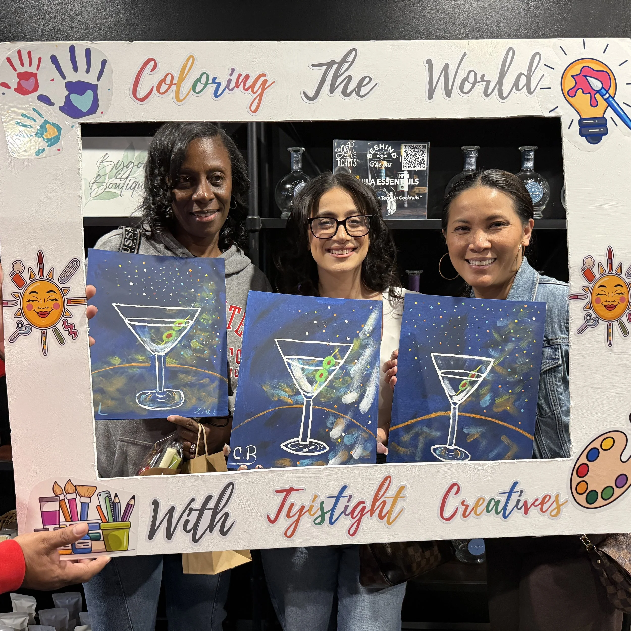 Three women are posing behind a large colorful sign that says 'Coloring The World With Jystight Creations' with decorative stickers and drawings of art supplies and a sun with a smiling face.
