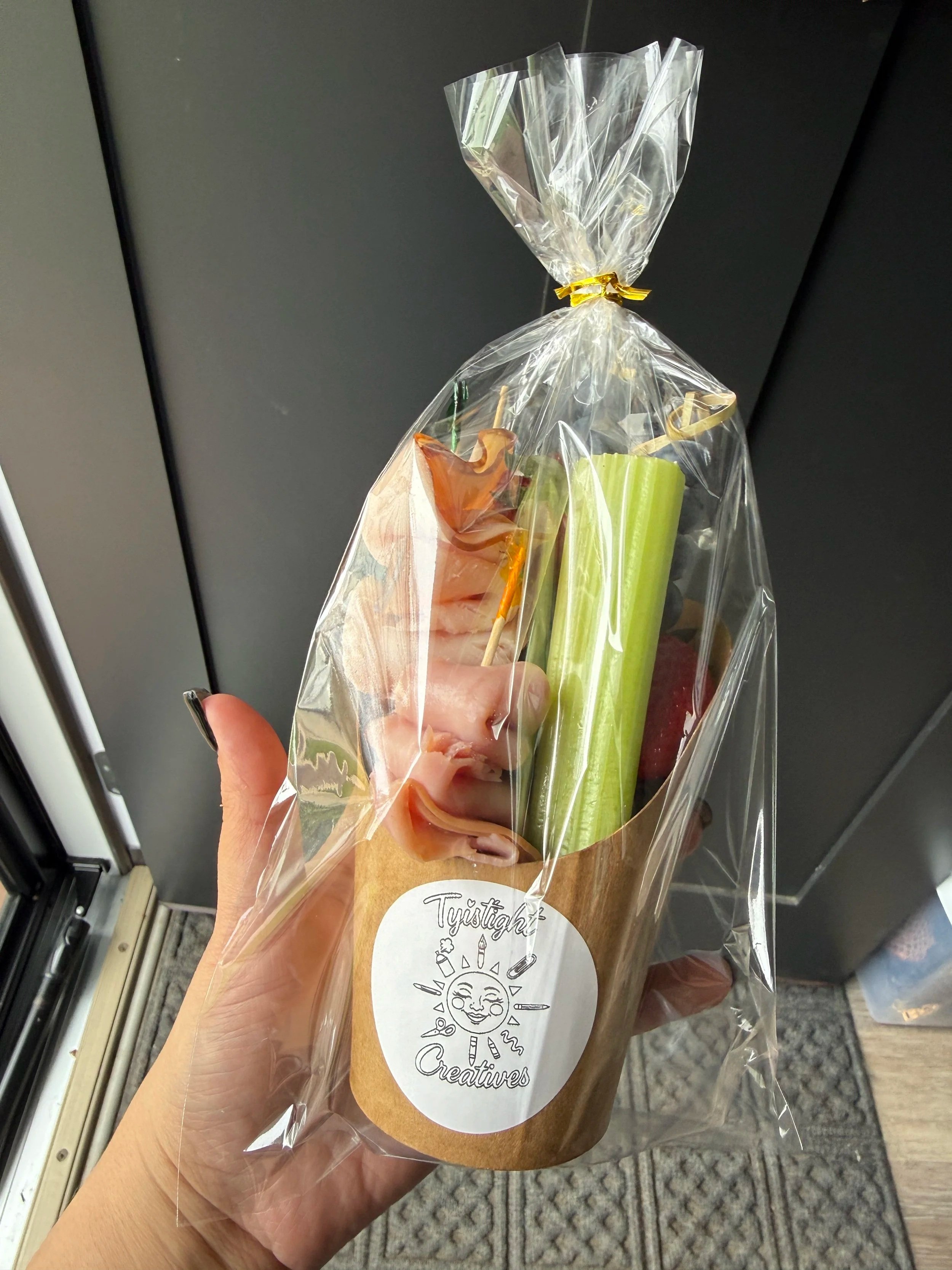 A hand holding a wrapped snack with a round label that says 'Twitch Creatives' featuring a sun with a smiling face and various icons. The snack includes a piece of celery, a skewer with meat or cheese, and other sliced vegetables, wrapped in clear plastic.