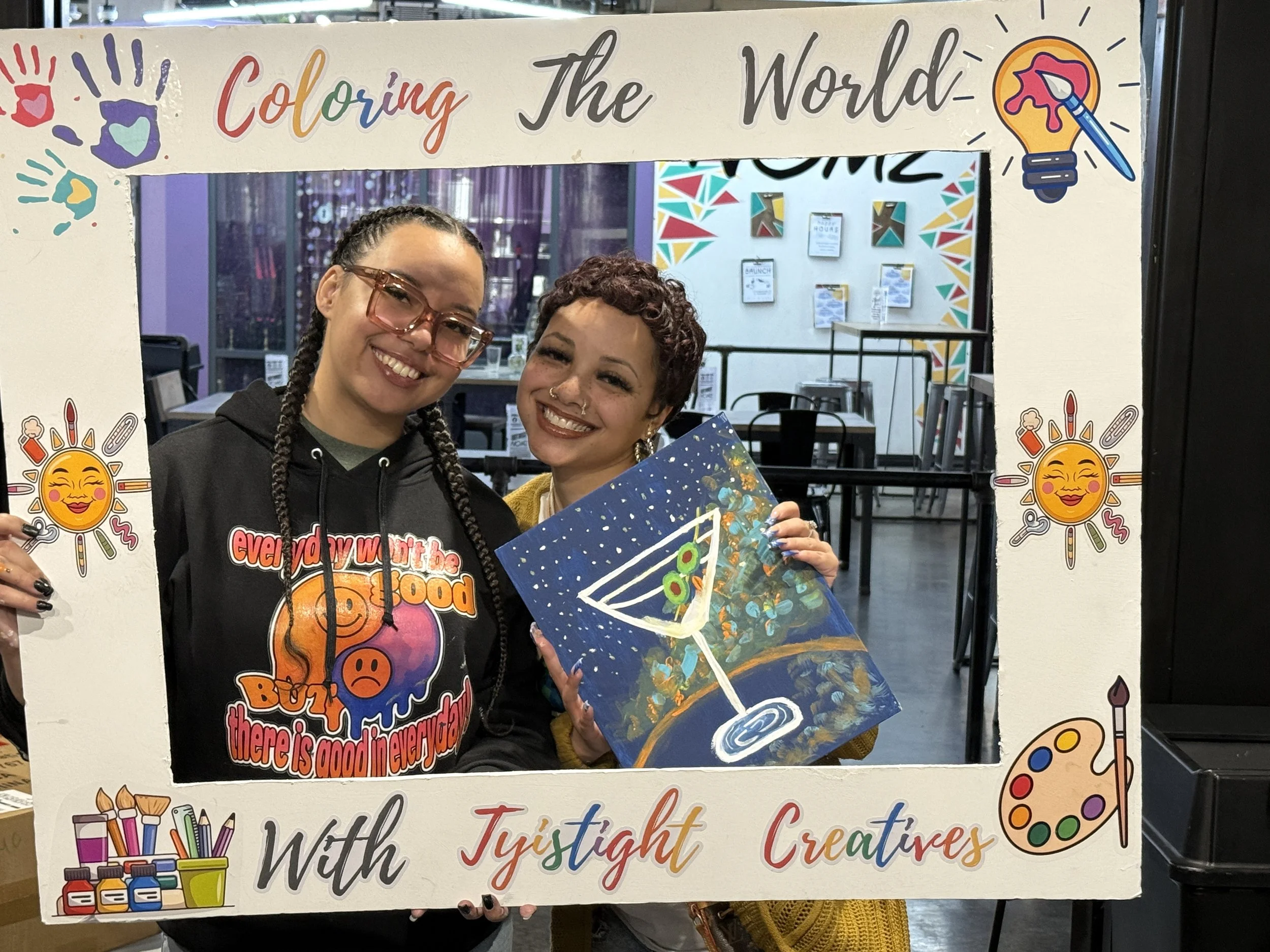 Two women smiling, holding a colorful painting of an abstract design with a swirl and circles, and standing behind a frame with the words "Coloring The World" as part of a creative-themed display. The frame is decorated with colorful handprints, suns, and art supplies.