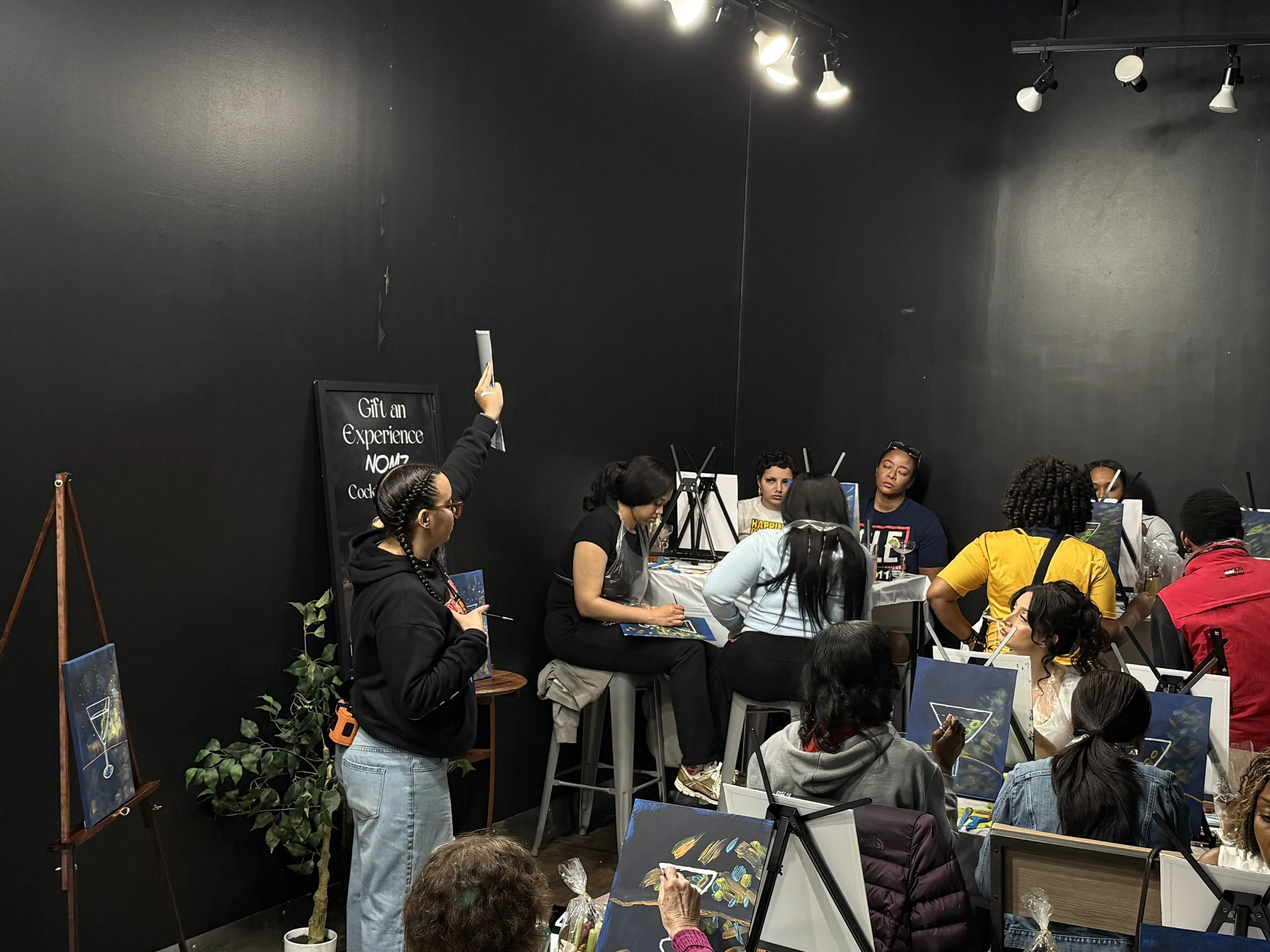 A group of people participating in a painting class in a dark room with black walls, illuminated by spotlights. Some are standing, others are seated, and there are several artworks and painting supplies around.