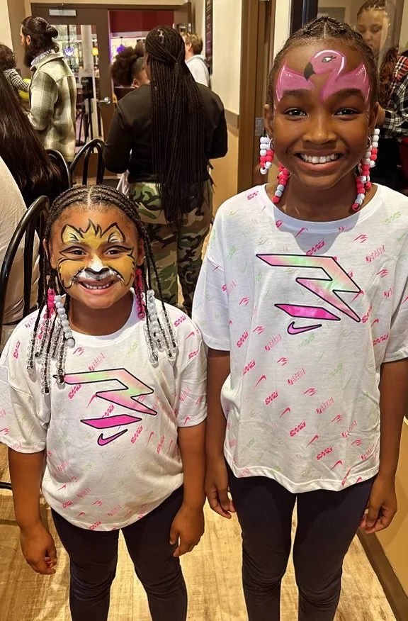 Two young girls with face paint, smiling at the camera, wearing matching white shirts with colorful designs, inside a busy indoor setting.
