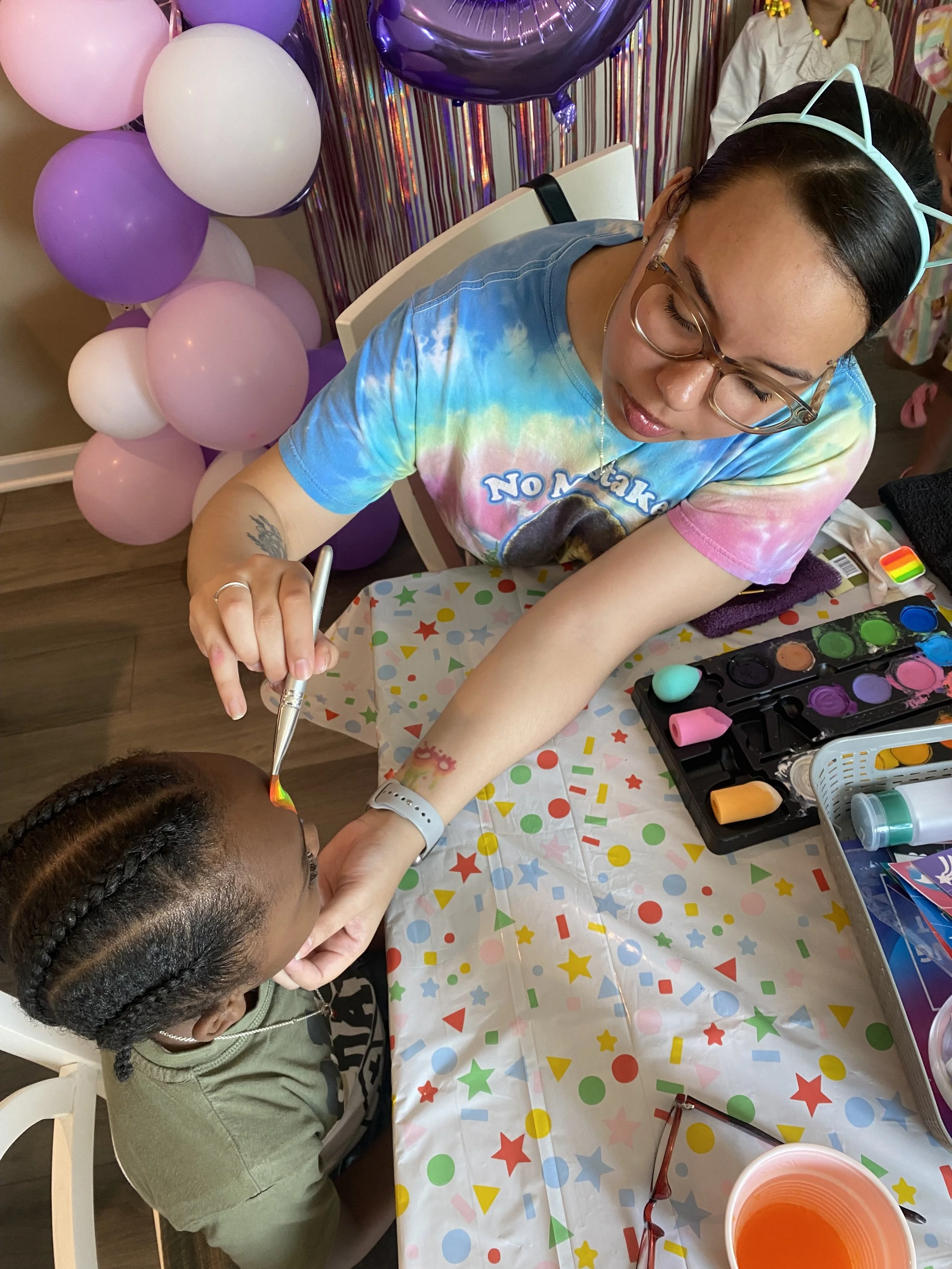 A woman wearing glasses and a cat ear headband is painting a child's fingernails at a birthday party. The table is decorated with a colorful star and circle patterned tablecloth, and there are balloons in purple, pink, and white in the background. The woman is wearing a tie-dye T-shirt, and the child has braided hair and is wearing a green jacket.
