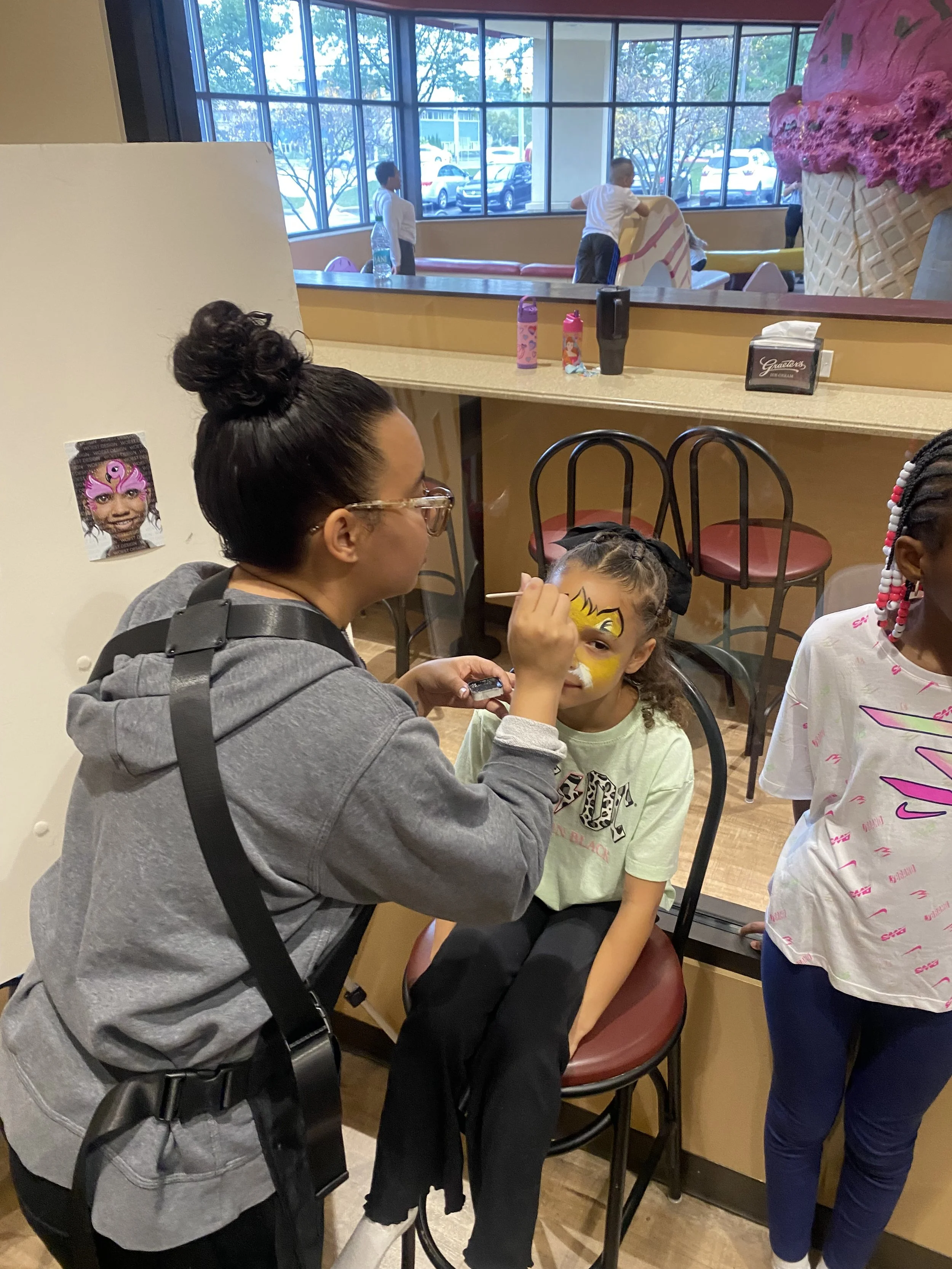 A woman in gray hoodie and glasses face painting a young girl with curly hair, wearing a light green shirt, in a restaurant or cafe with a counter and large window in the background. Other children are present, and a poster of a girl with face paint is on the wall.