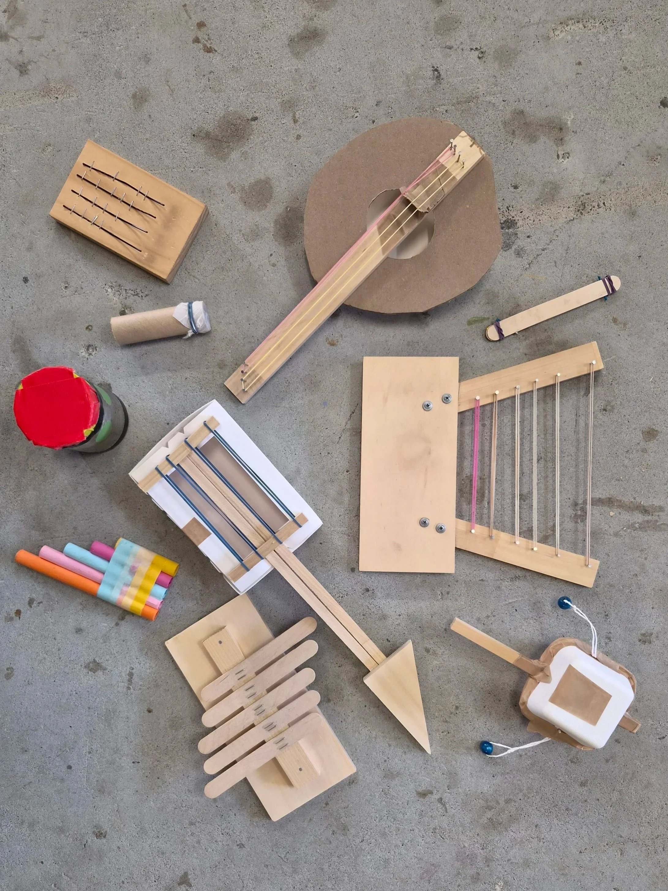 Arrangement of various handmade musical instrument projects made from cardboard and wood, including a guitar shape, a xylophone, and percussion instruments, displayed on a flat surface.