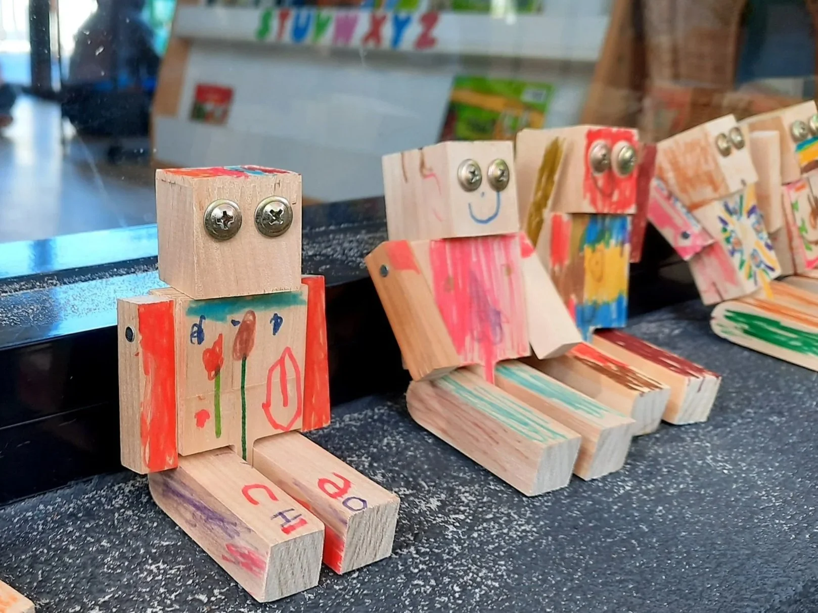Wooden blocks with colorful drawings and metallic screws arranged to resemble small human figures, displayed on a black surface.