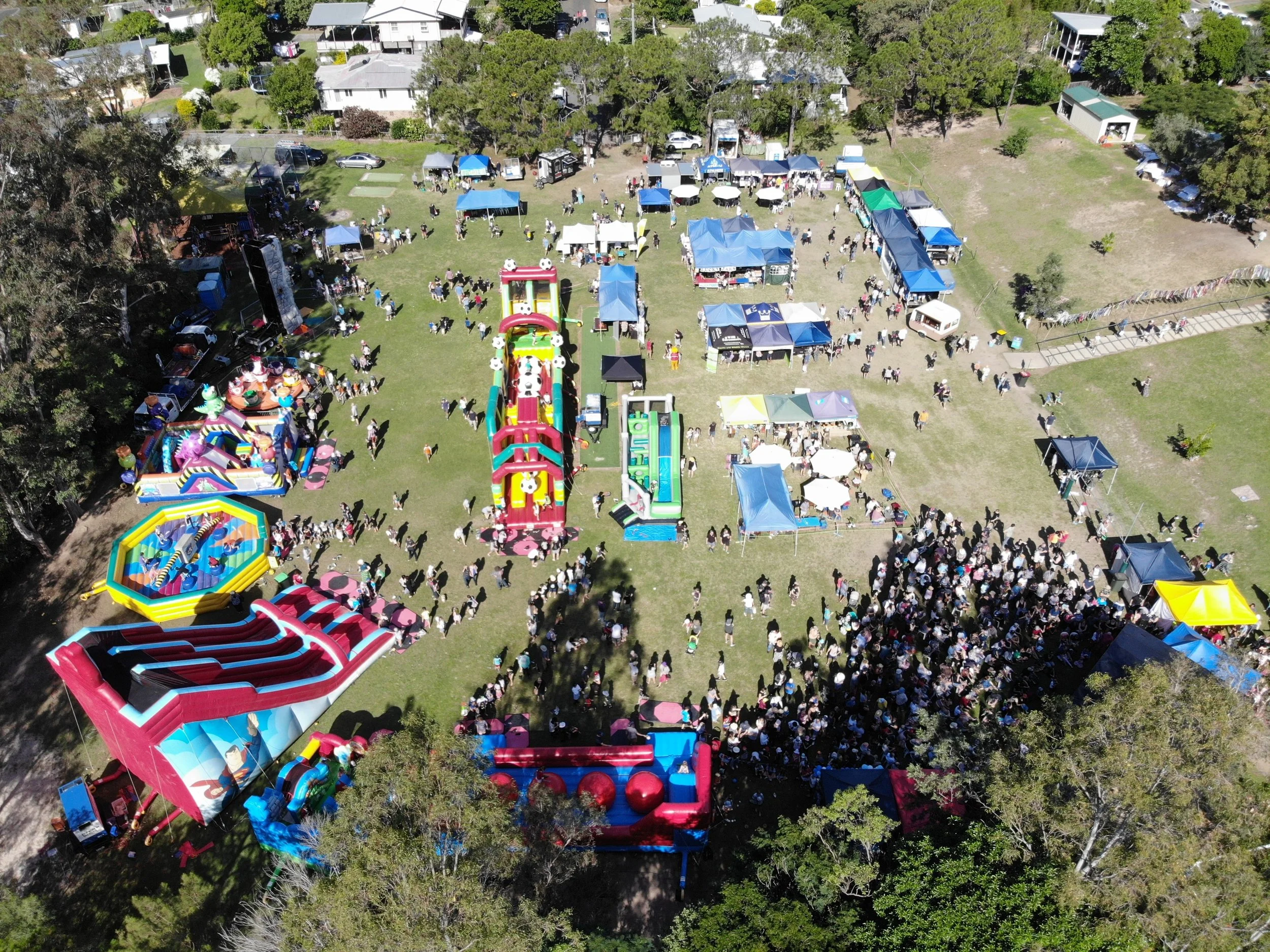 A lively outdoor fair with numerous colorful inflatable bounce houses, tents, and booths on a grassy field. Many people are walking around, and there are trees and residential houses in the background.