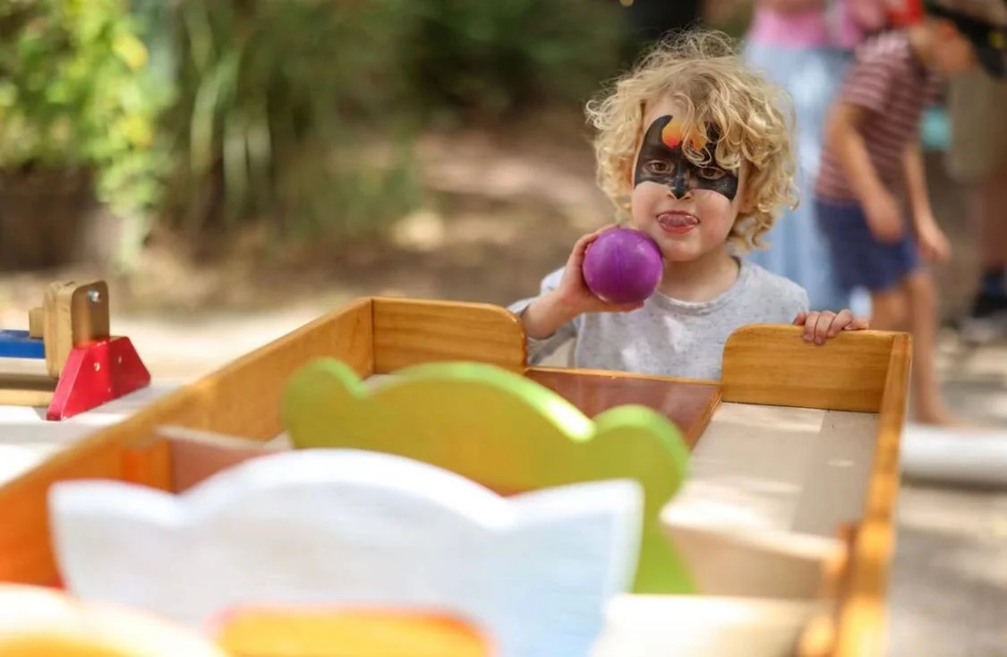A young child with curly blonde hair and a face painted like a dragon, holding a purple ball, standing behind a wooden game table outdoors during daytime.