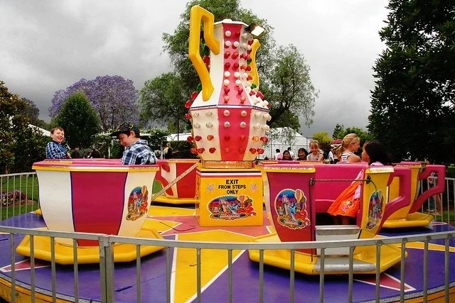 Children enjoying a colorful teacup ride at an amusement park with trees and cloudy sky in the background.