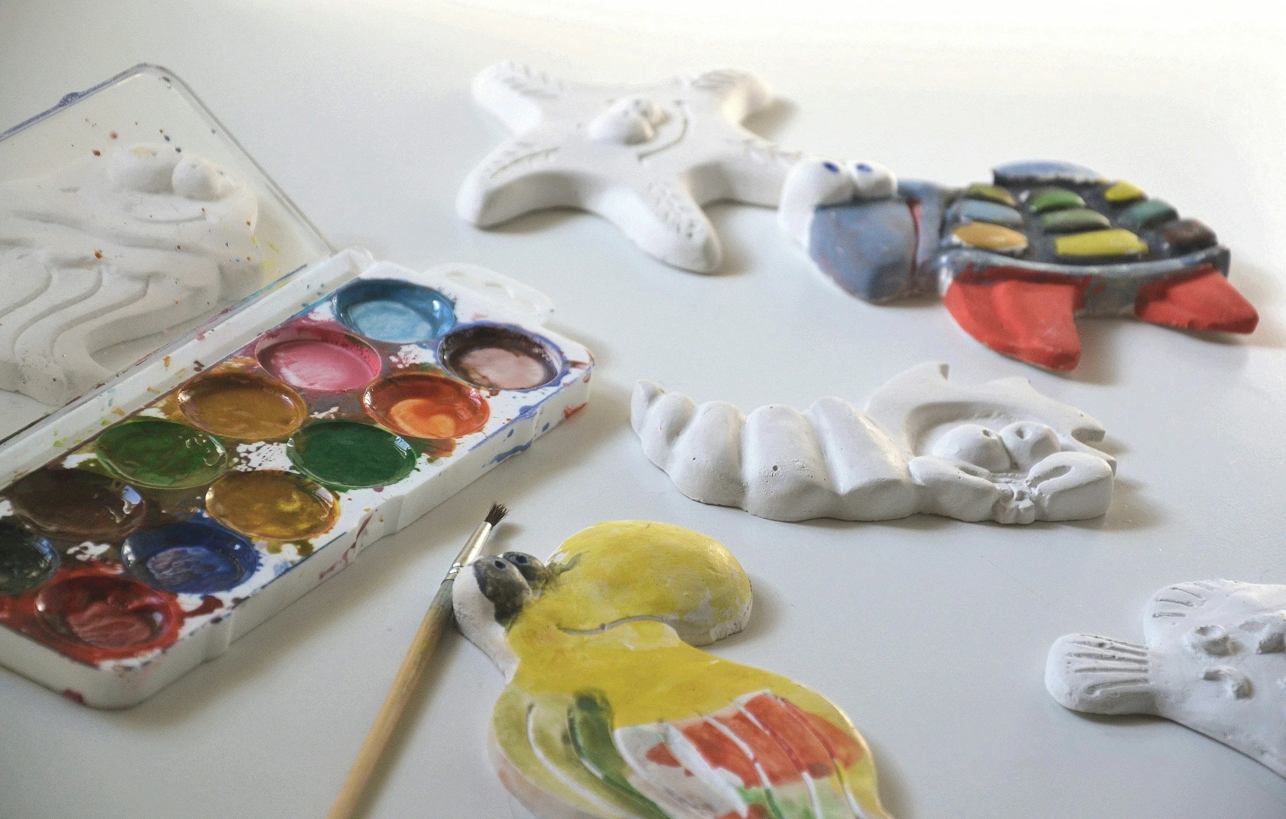 Colorful painted ceramic figurines, including a yellow duck and other animal shapes, next to watercolor palette with various colors and brushes on a white surface.