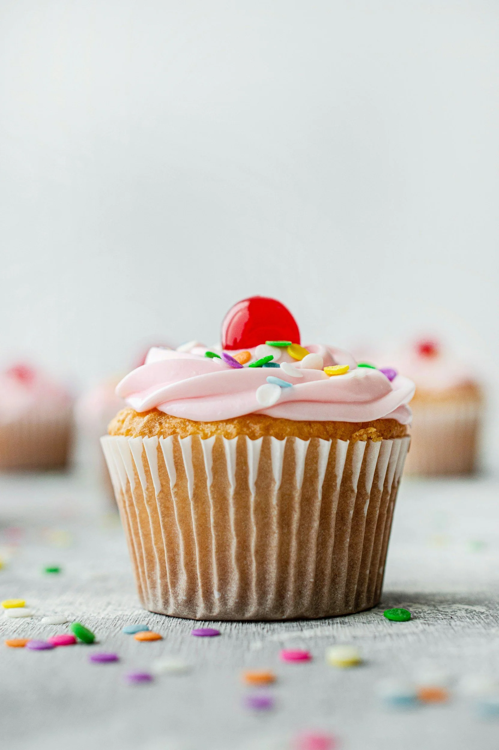 A cupcake with pink frosting, colorful sprinkles, and a red cherry on top. The cupcake is in a brown paper wrapper, and there are scattered sprinkles on a white surface around it.