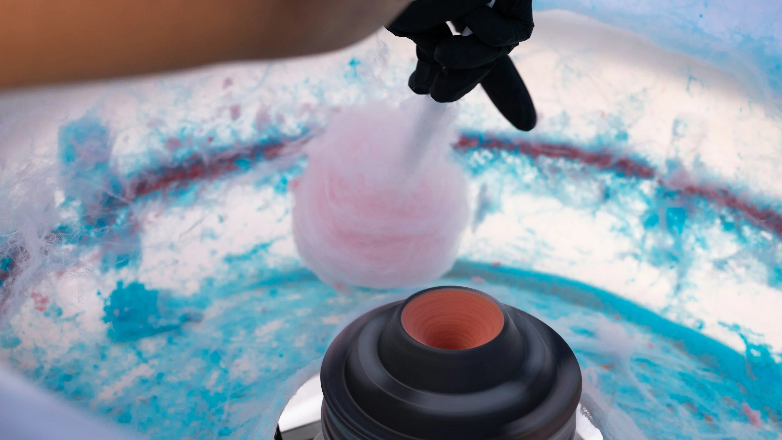 Close-up of a person creating a pink cotton candy on a stick with a cotton candy machine, surrounded by blue and white fluffy cotton candy.