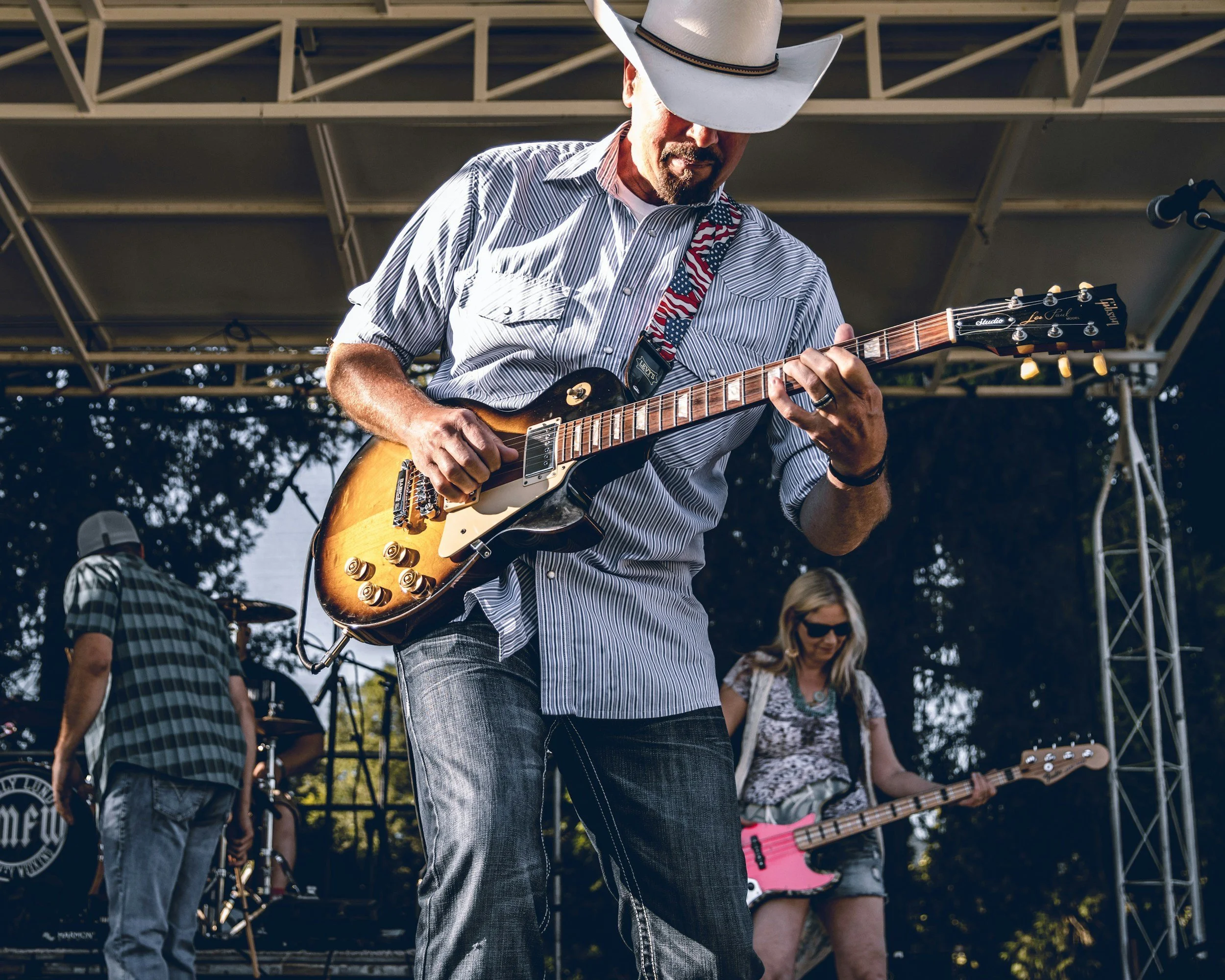 A man wearing a cowboy hat and striped shirt playing an electric guitar during an outdoor concert, with other musicians and audience visible in the background.