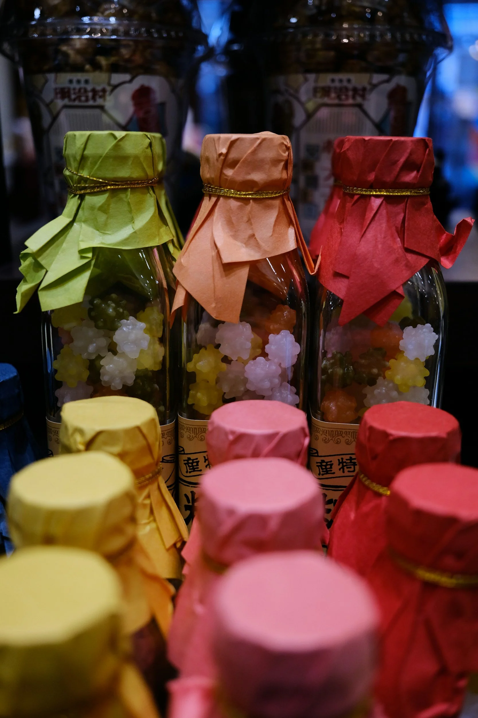 Three glass bottles filled with colorful rock candies, each covered with crumpled paper and tied with a thin string. Background includes more bottles with similar packaging.