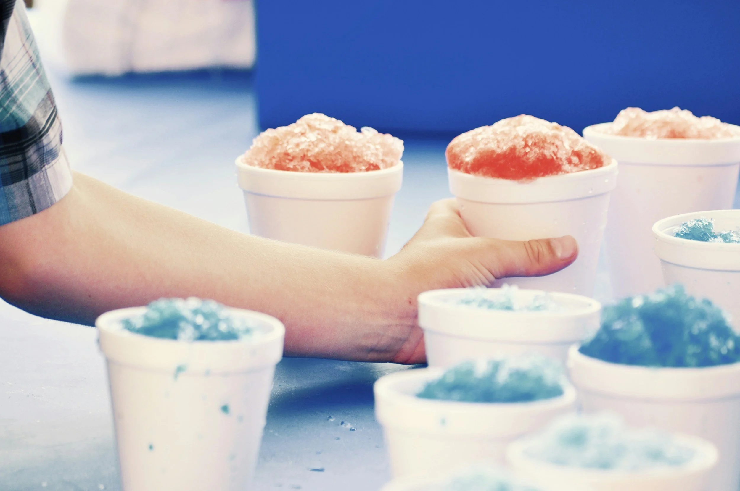 A person holding a cup of pink shaved ice dessert, with similar cups of shaved ice in the background.