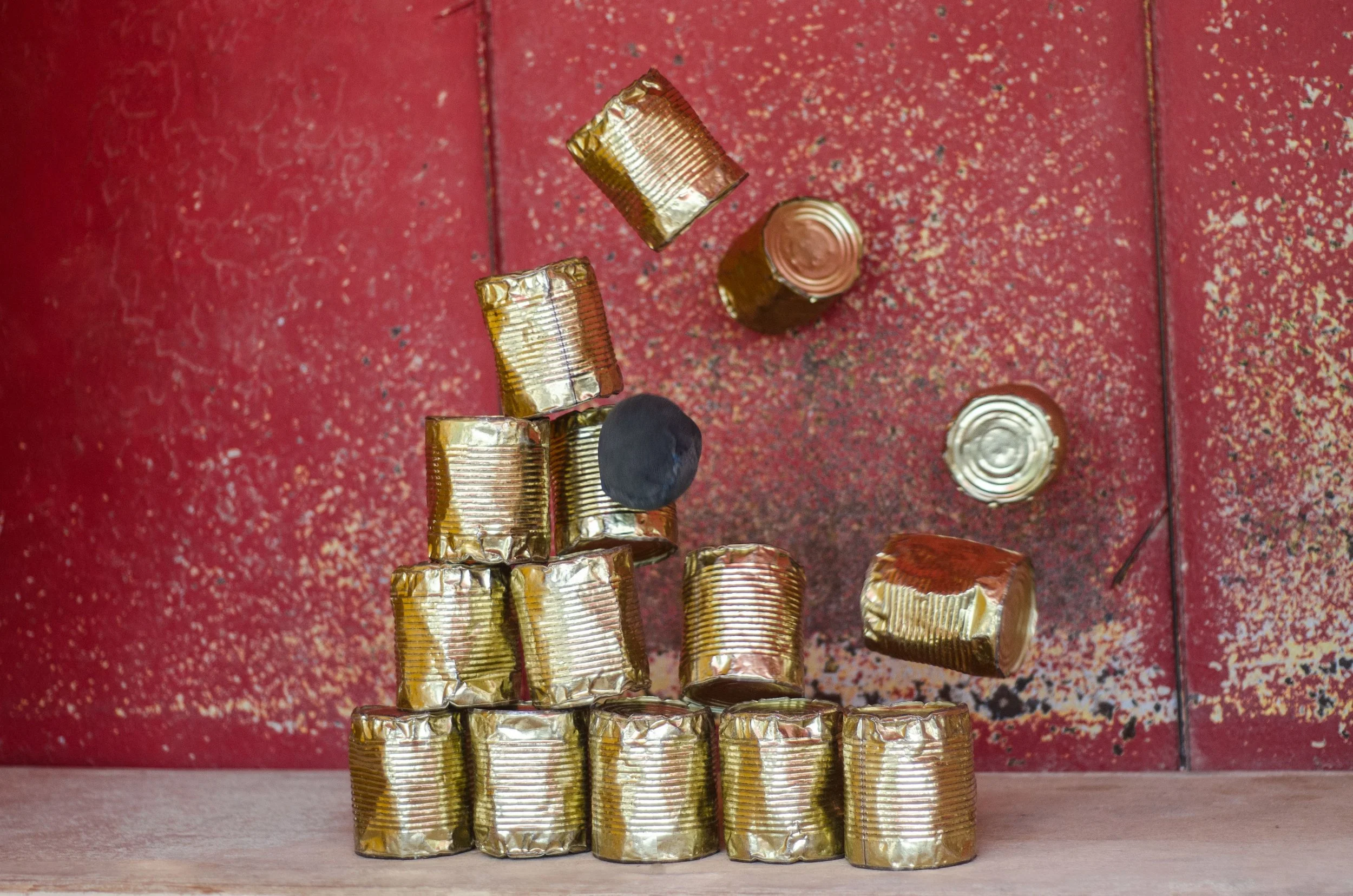 Stacked gold-colored tin cans arranged in a pyramid shape on a wooden surface, with a red textured wall in the background.