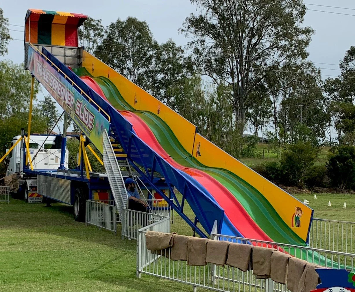 Colorful funhouse slide, featuring multiple lanes purple, yellow, red, and green, set outdoors with trees and grassy field in background.