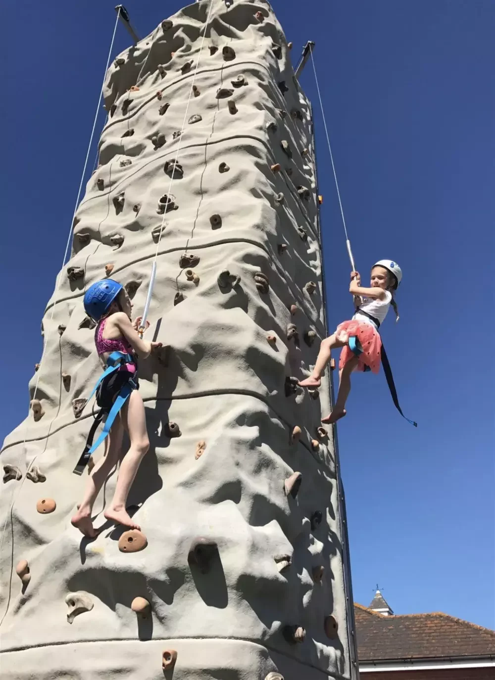 Two young girls wearing helmets are climbing an outdoor rock wall with various holds, set against a bright blue sky.