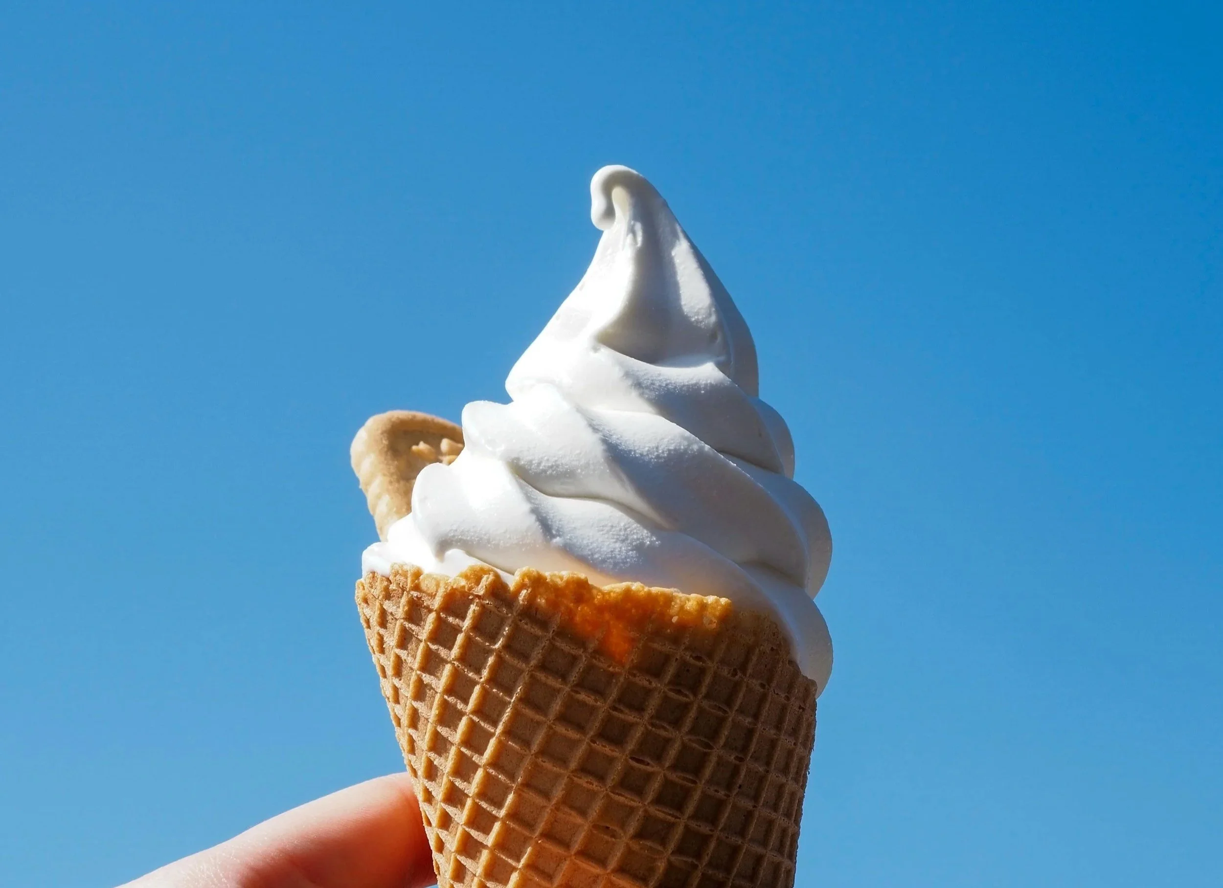 A hand holding an ice cream cone with soft serve ice cream in a cone, against a clear blue sky.