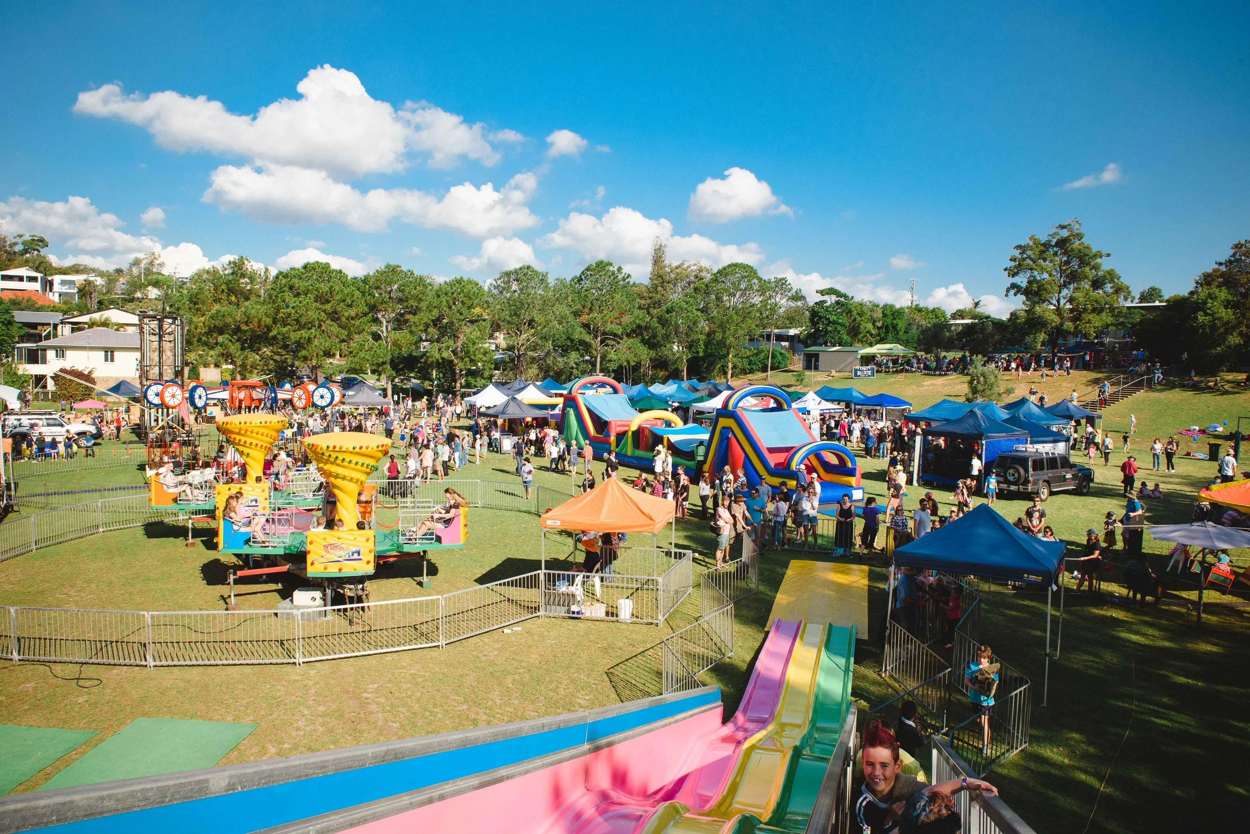 A lively outdoor fair or carnival on a sunny day with colorful inflatables, rides, tents, and many people enjoying the event, set against a backdrop of trees and blue sky with scattered clouds.