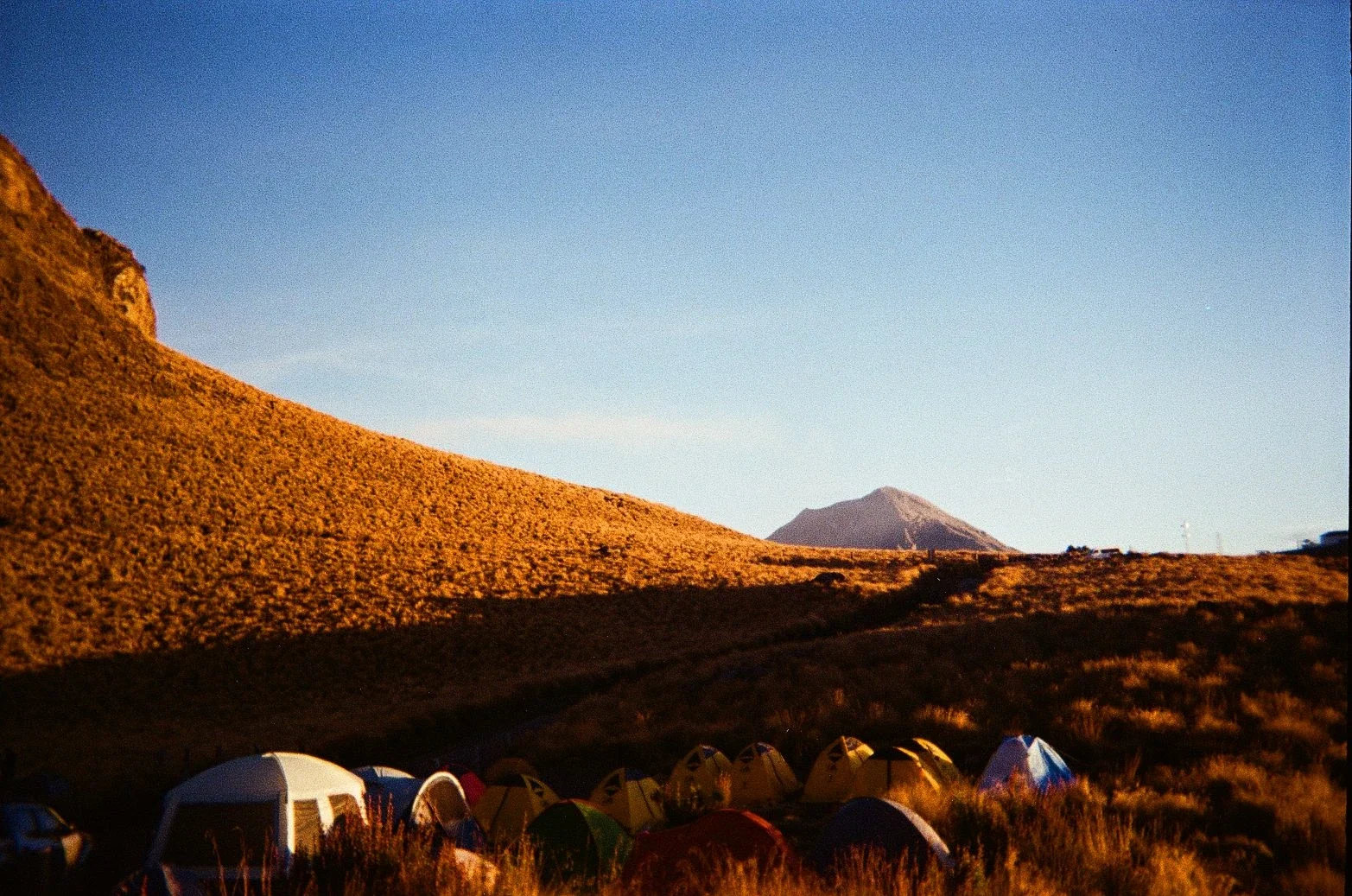 A mountainside campground with various tents set up on grassy terrain, with a large hill and distant mountain in the background, under a clear blue sky.