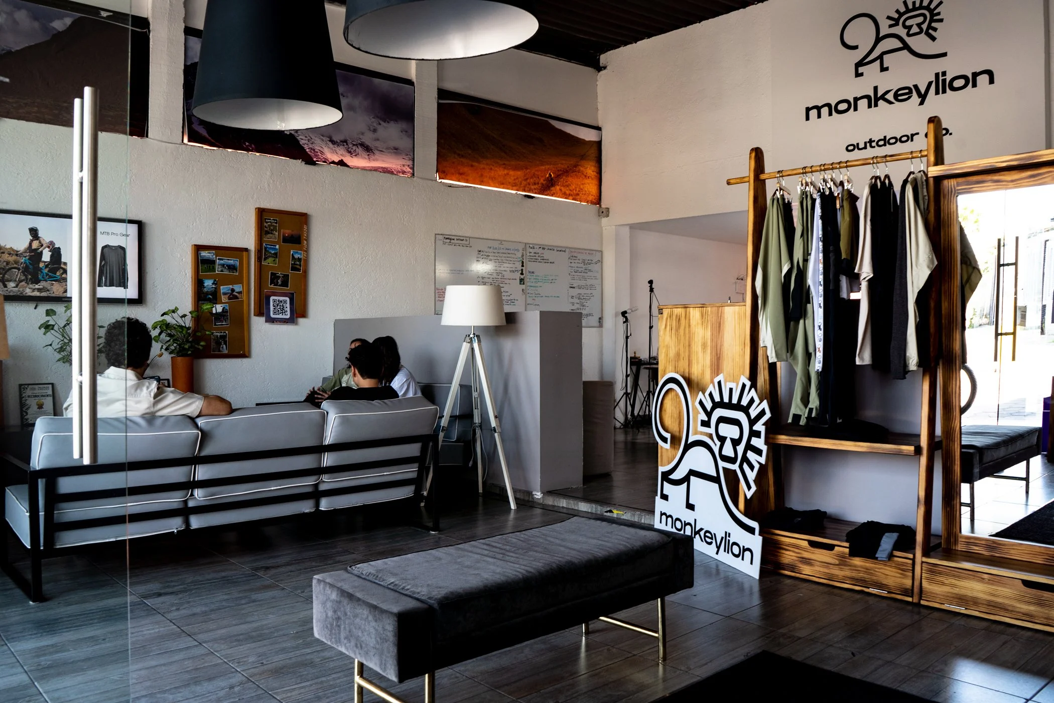 Inside a retail store with a seating area featuring a silver couch, a black and silver bench, and various signs and displays including a MonkeyLion logo, clothing rack, and a whiteboard with notes. Two people are seated on the couch, and there are decorative wall art and photographs.