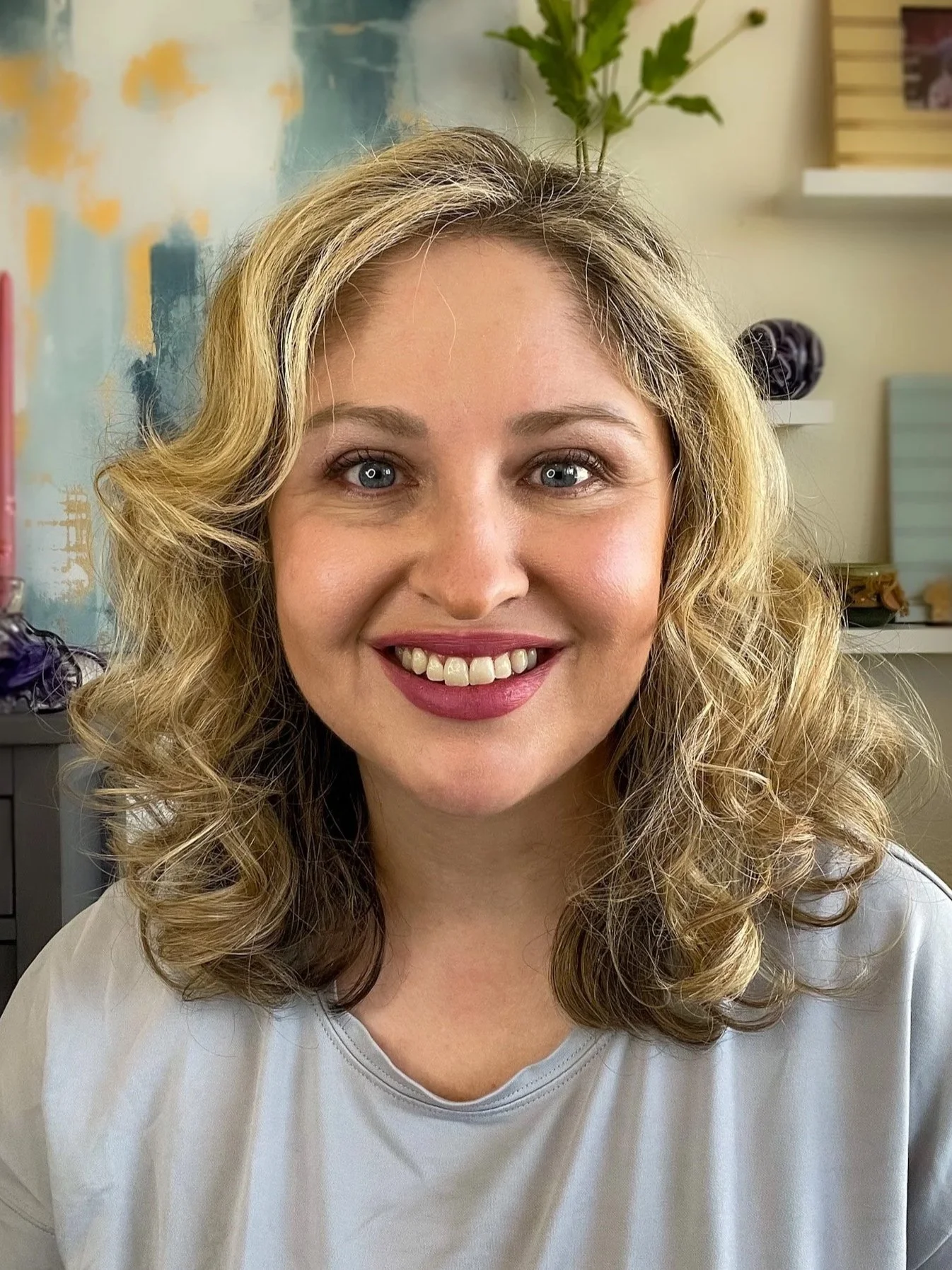 A woman with blonde, curly hair and bright blue eyes she is smiling and wearing a light-colored top the background features a painted wall, shelves with decorative objects, and a picture frame.