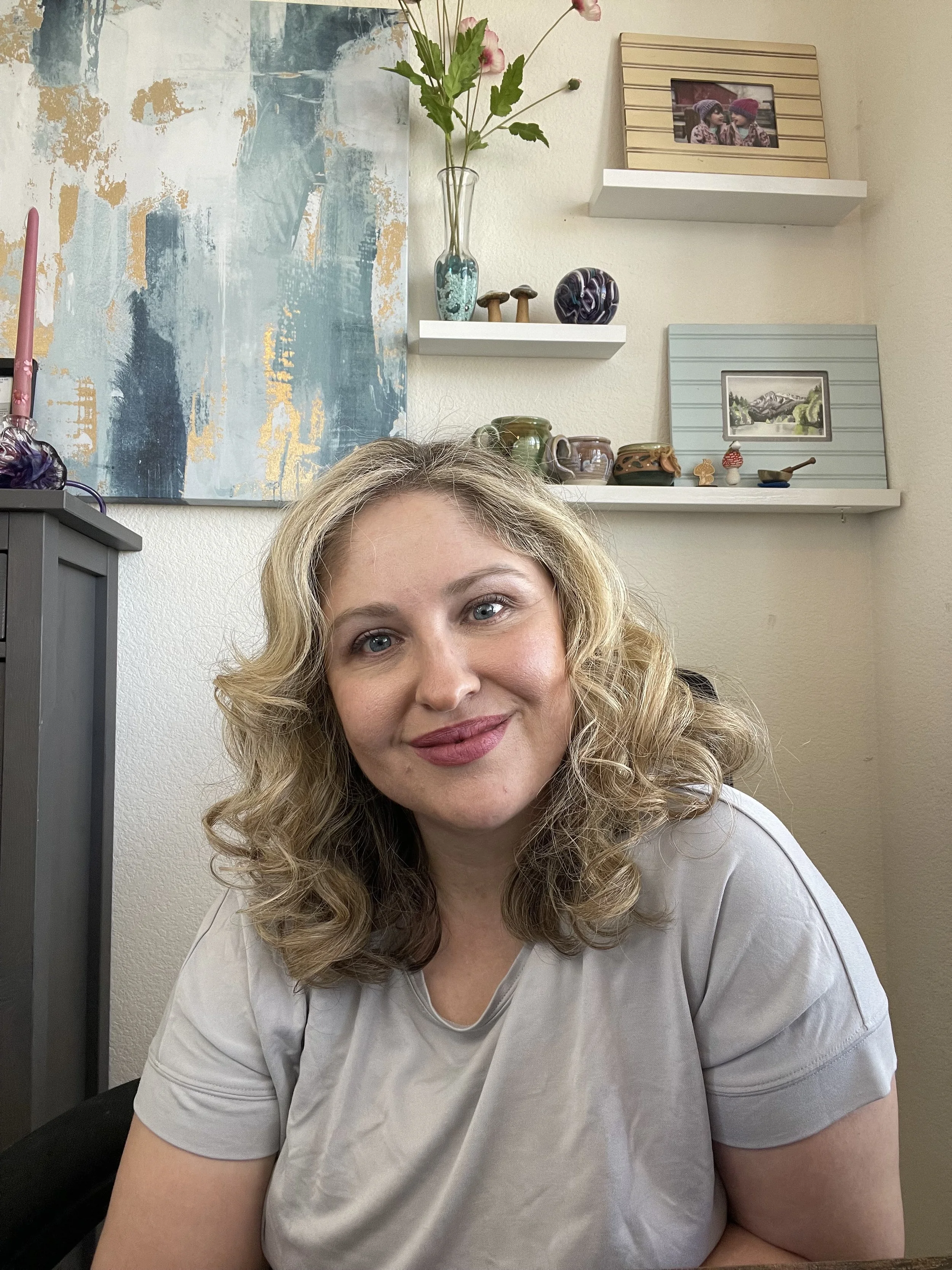 A woman with shoulder-length curly blonde hair and blue eyes smiling at the camera, sitting in a room with beige walls and decorative shelves holding framed photos, pottery, and a glass vase with pink flowers.