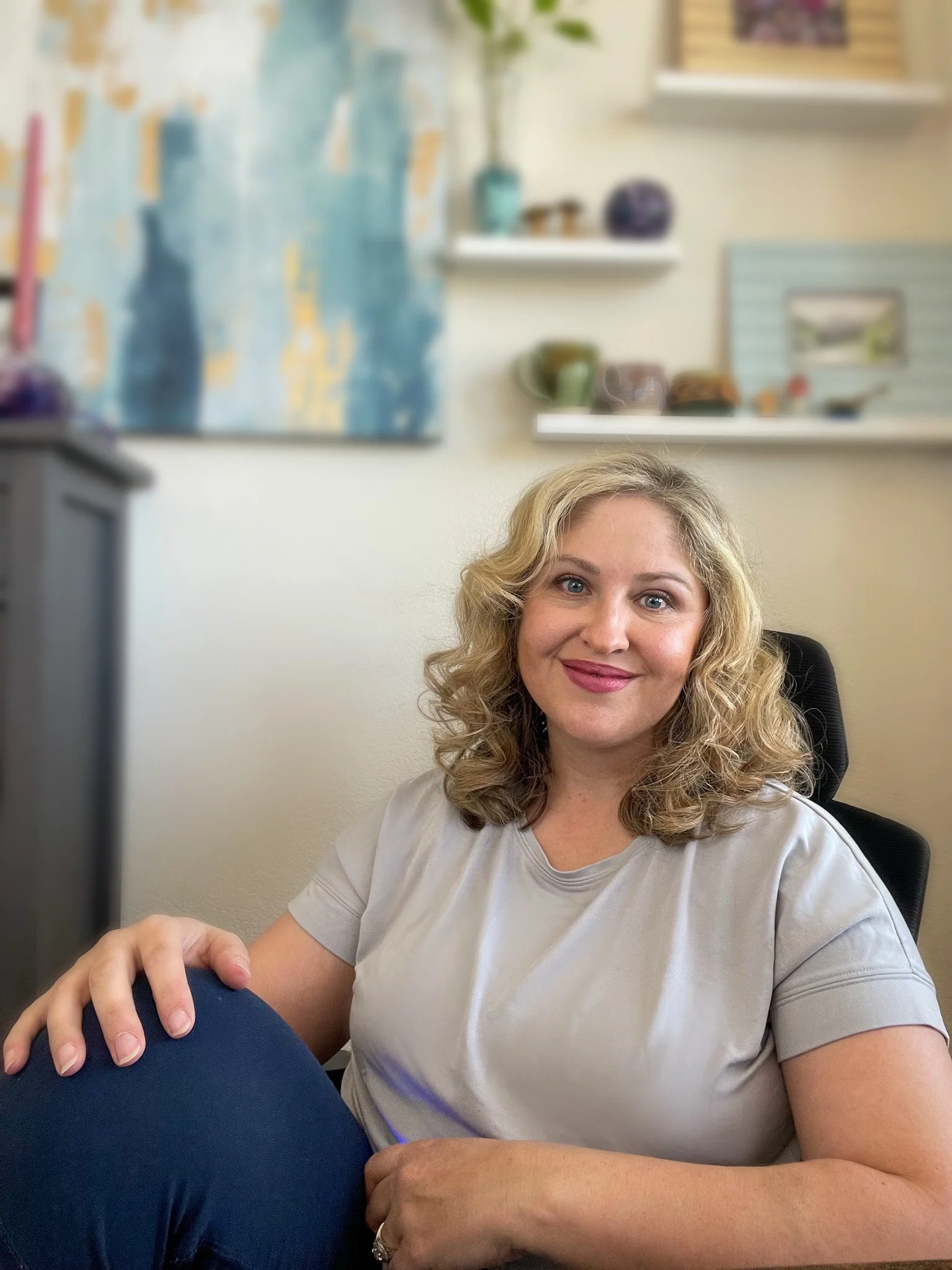 A smiling woman with blonde curly hair sitting in a black office chair in a well-decorated room with abstract artwork, shelves with decorative bowls, and potted plants.