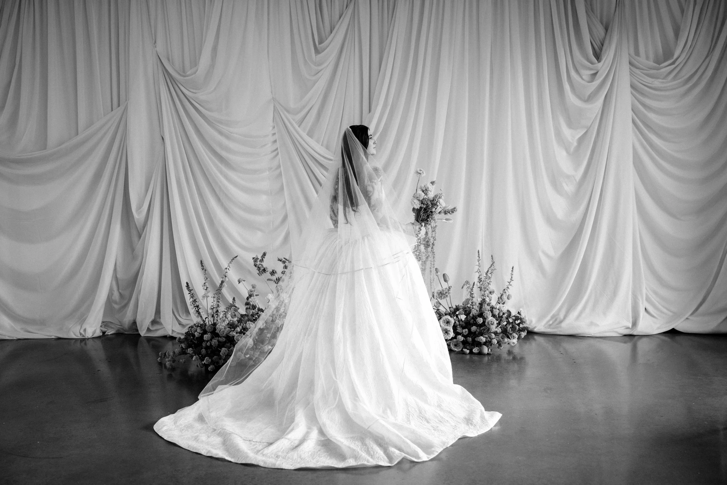 Black and white photo of a bride in a wedding dress and veil, holding a bouquet of flowers, standing in front of a large draped curtain backdrop decorated with flowers at the base.