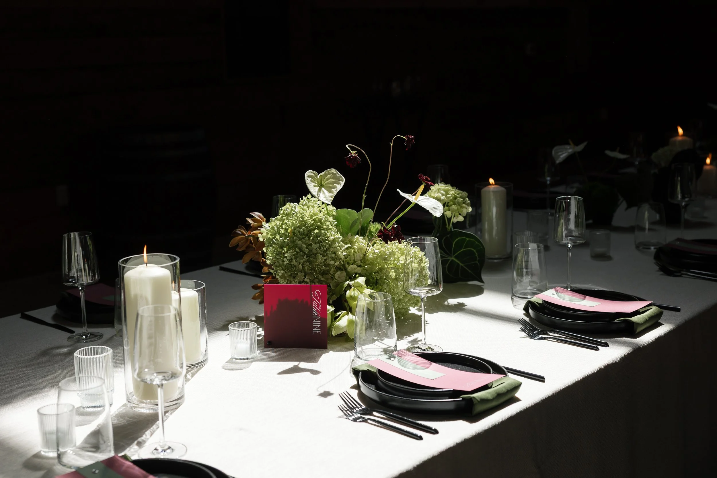 A decorated dinner table with a floral centerpiece, surrounded by lit candles, wine glasses, and place settings, in a dimly lit room.