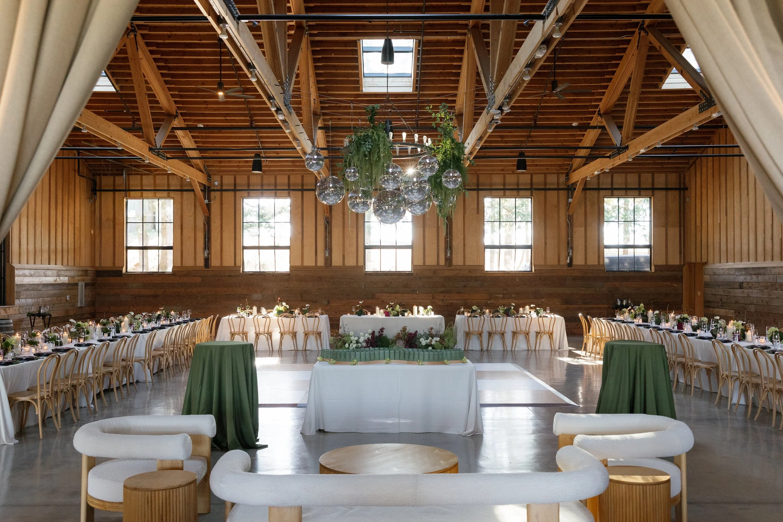 A spacious wooden hall decorated for an event, with a high ceiling, large windows, round tables with white tablecloths, and a floral centerpiece in the middle. There are green and white chairs, a chandelier with hanging glass orbs, and a rustic aesthetic.